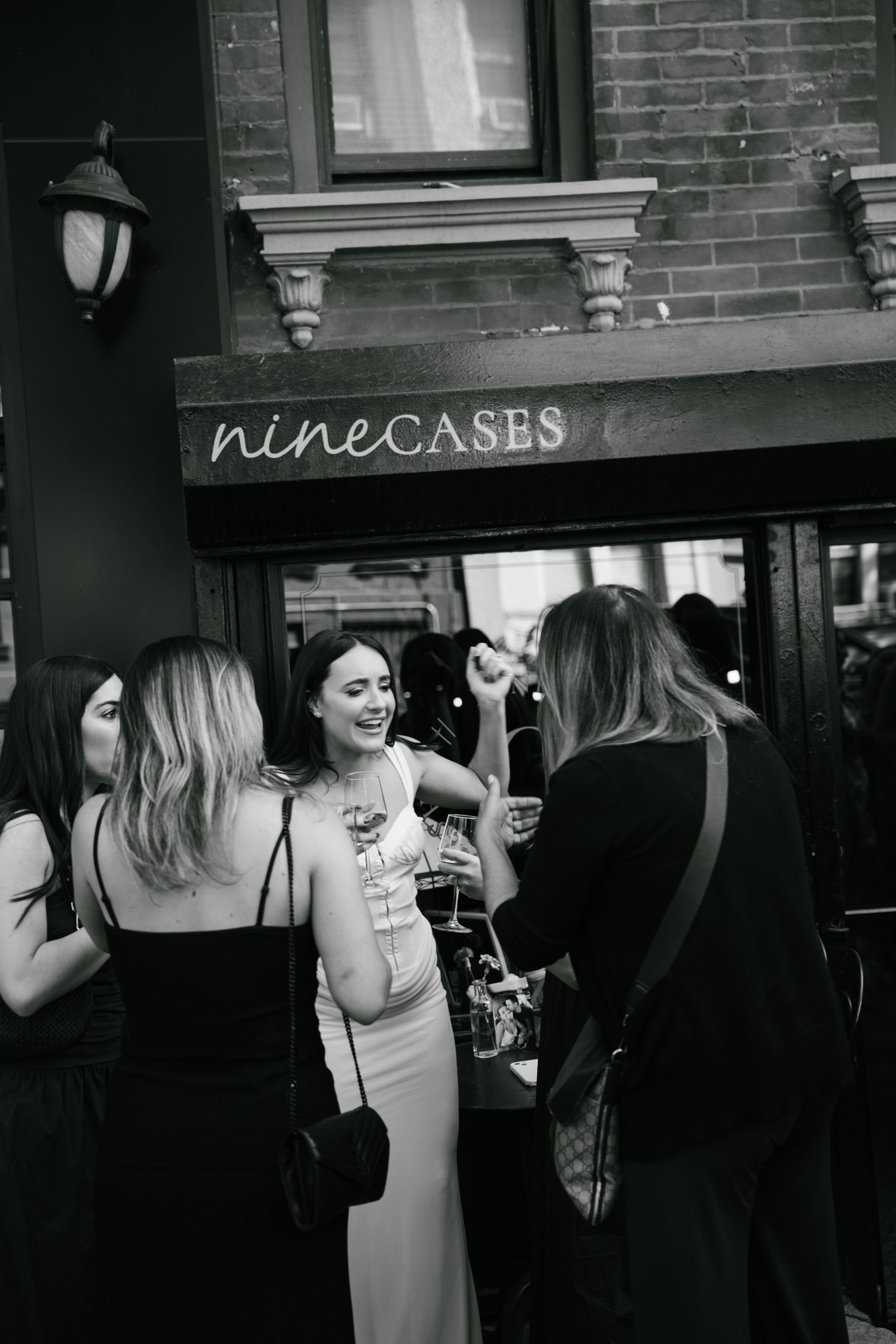 Group of women at a social gathering, holding drinks and engaging in conversation outside a building with brick walls, window ledge, and a sign that reads 'nine CASES'.