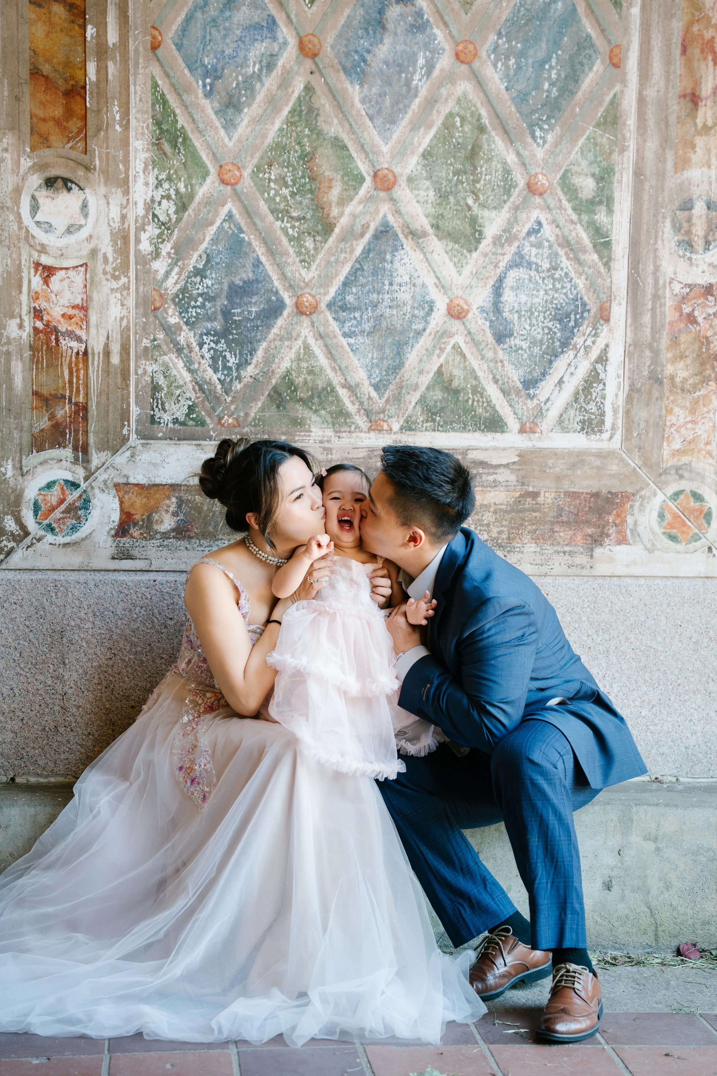 A family of three, including a woman, a man, and a child, dressed in wedding attire, sitting together and kissing in front of a decorative, painted wall with geometric patterns.
