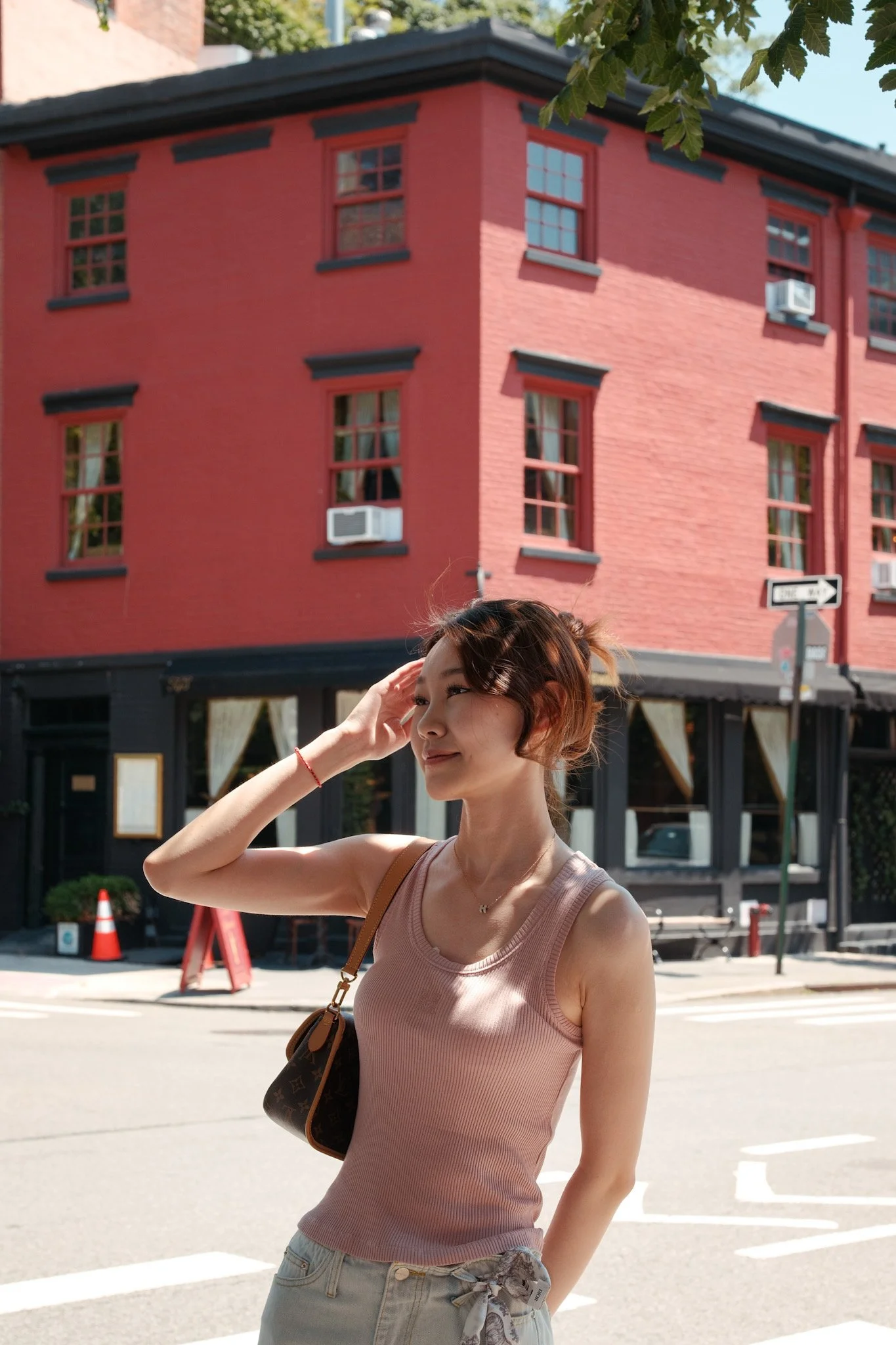 A woman stands on a city street in front of a pink brick building with multiple windows and air conditioning units. She is wearing a sleeveless flesh-colored top, light gray pants with a belt, and has a brown designer bag over her shoulder. She is sm