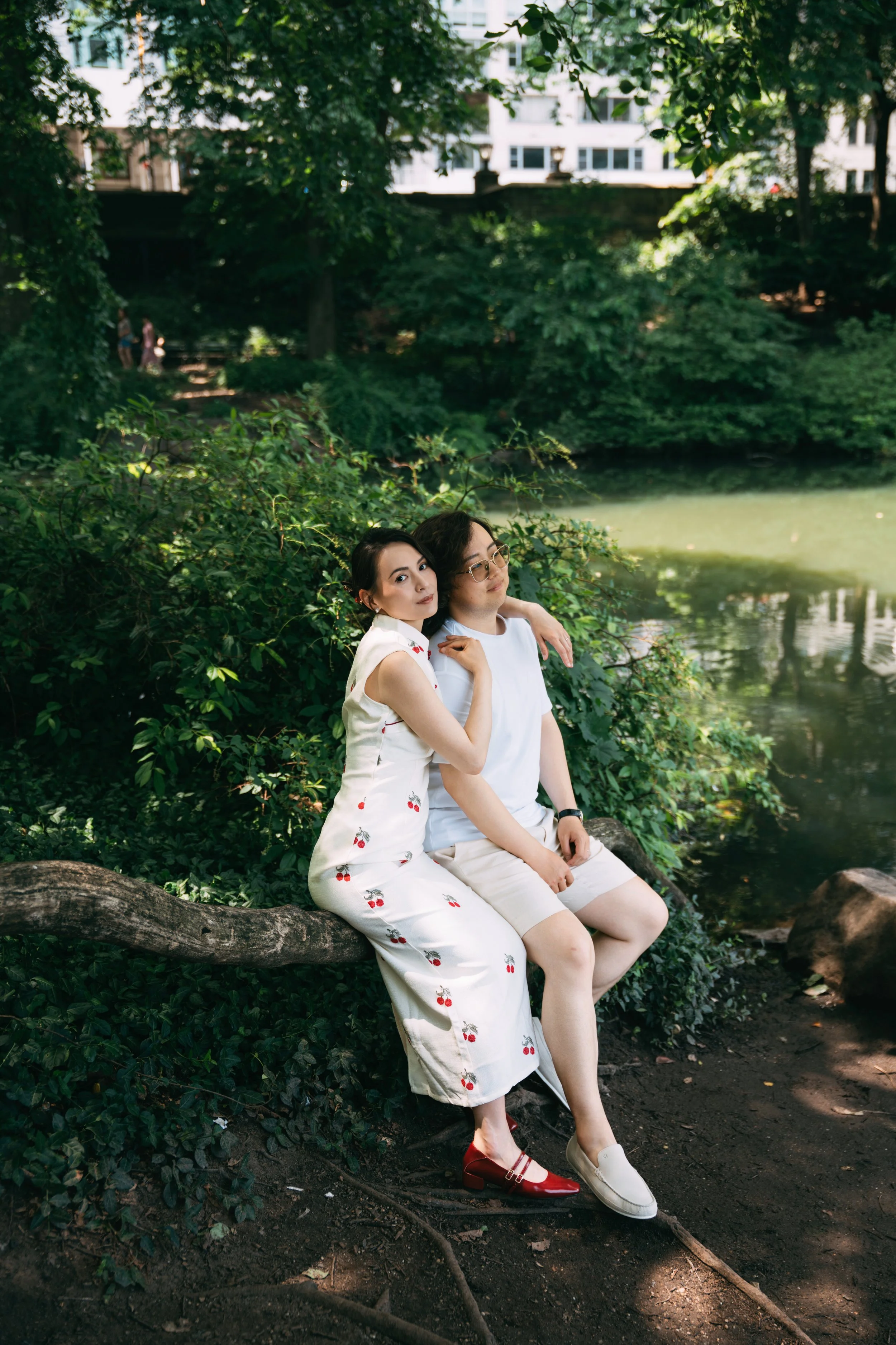 A young woman and man sitting on a log in a lush green park near a body of water, with trees and buildings in the background, during daytime.