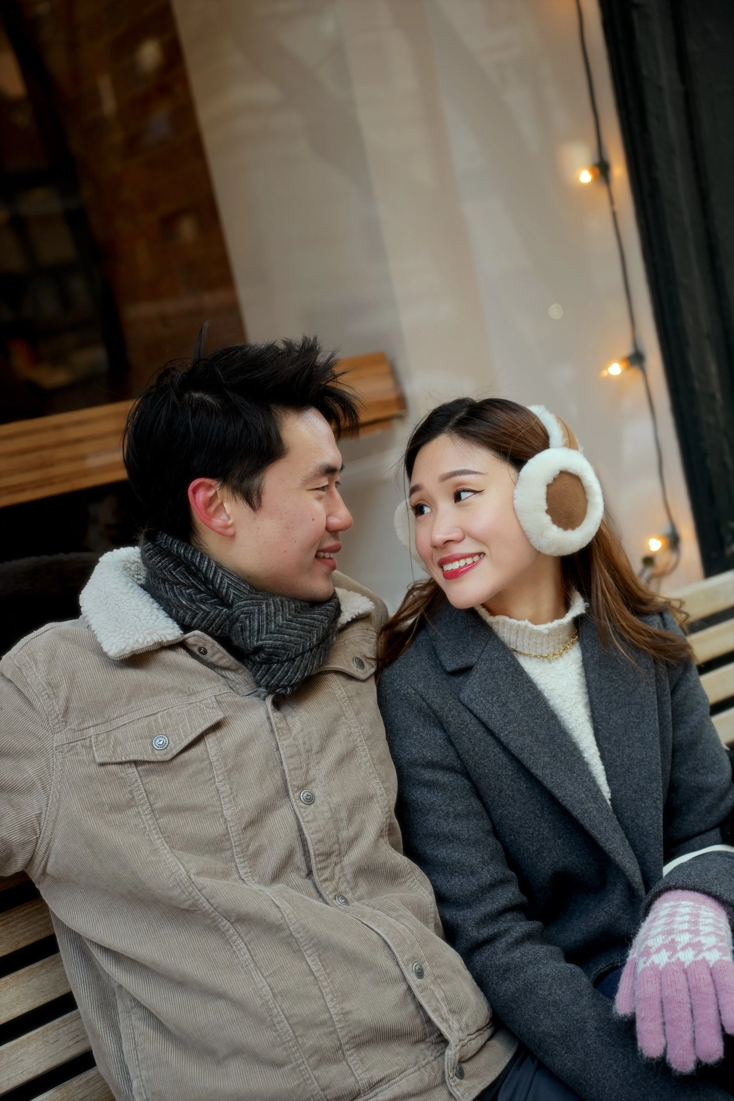 A young man and woman sitting close together, smiling and looking at each other inside a cozy, warmly lit room with string lights and wooden decor. The woman is wearing earmuffs and a gray coat, and the man is wearing a beige jacket and scarf.
