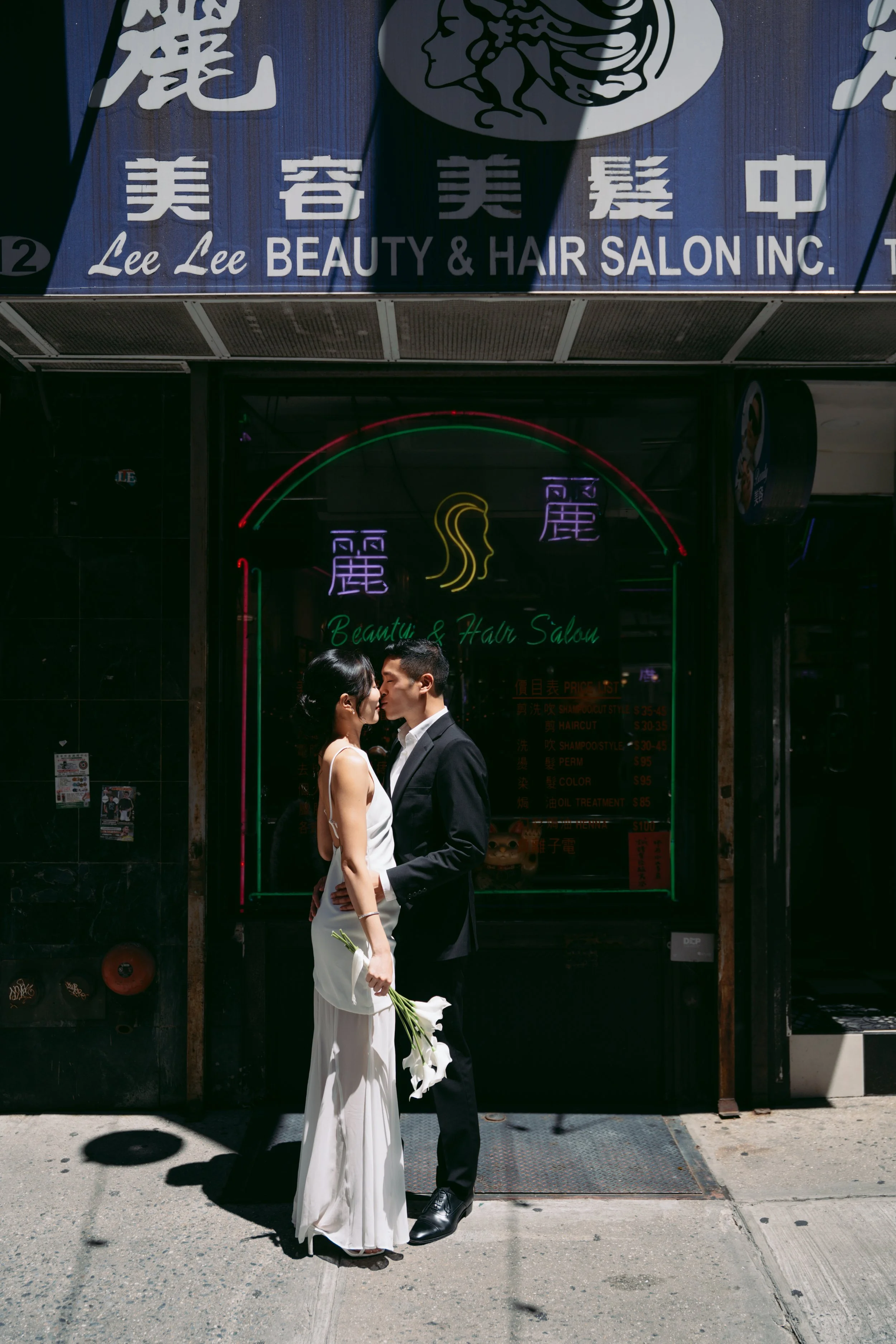 A bride and groom share a kiss outside a beauty and hair salon, with the bride holding a bouquet of white flowers.