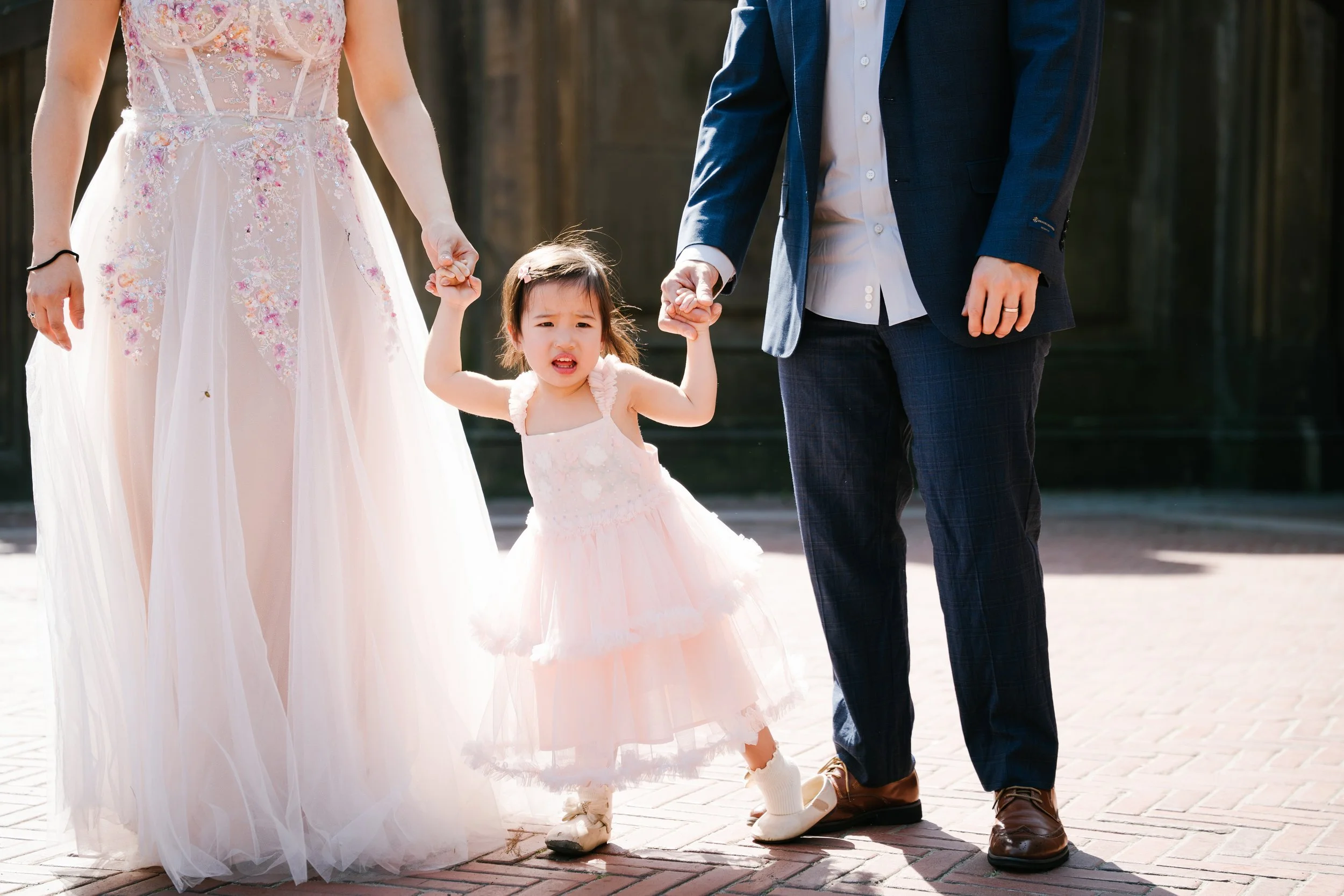 A young girl in a pink dress being supported by a woman in a embroidered wedding gown and a man in a blue suit, holding her hands and walking on a brick pavement outdoors.