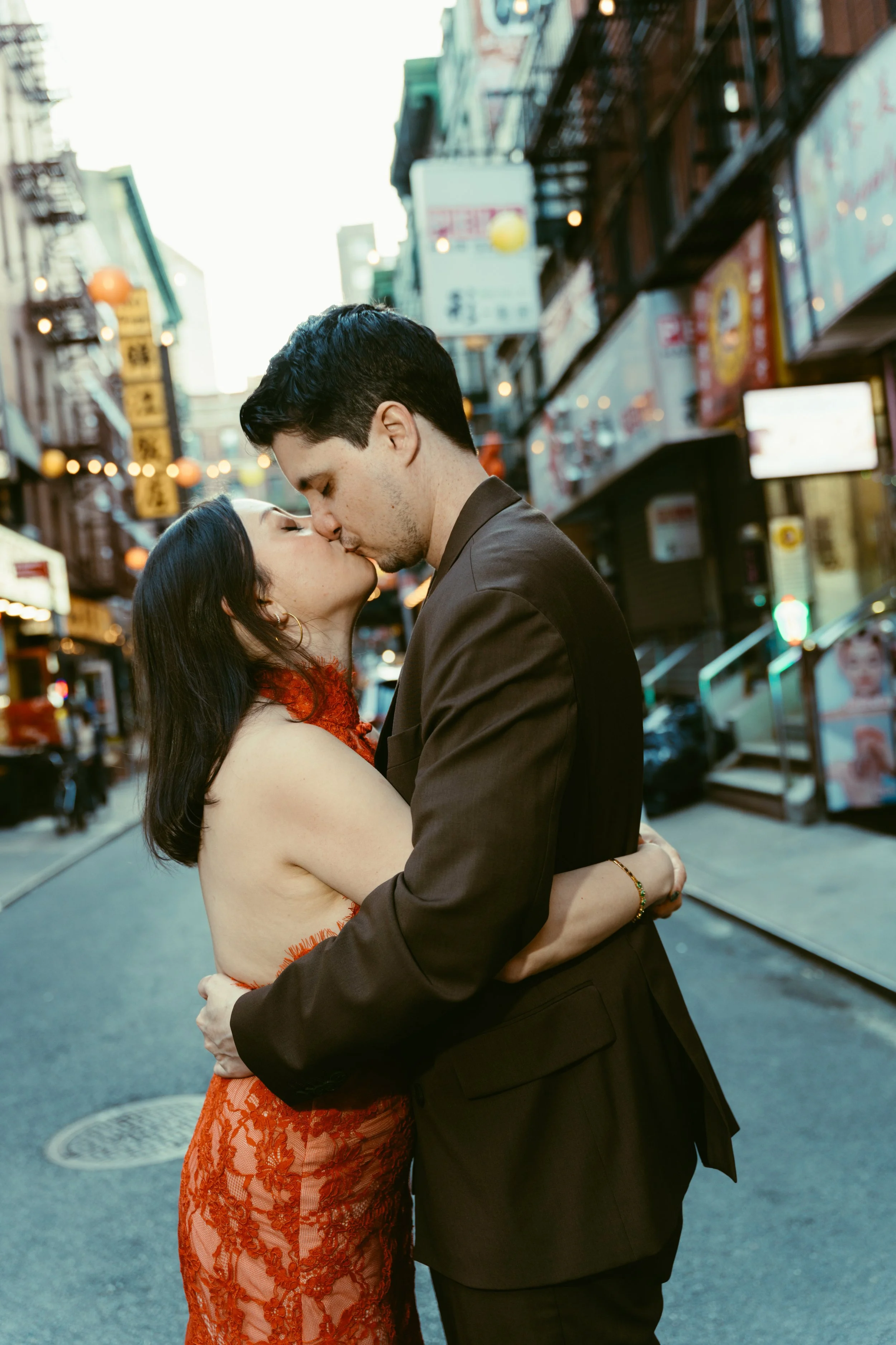 A couple in formal attire sharing a kiss on a city street at dusk, surrounded by illuminated signs and buildings.