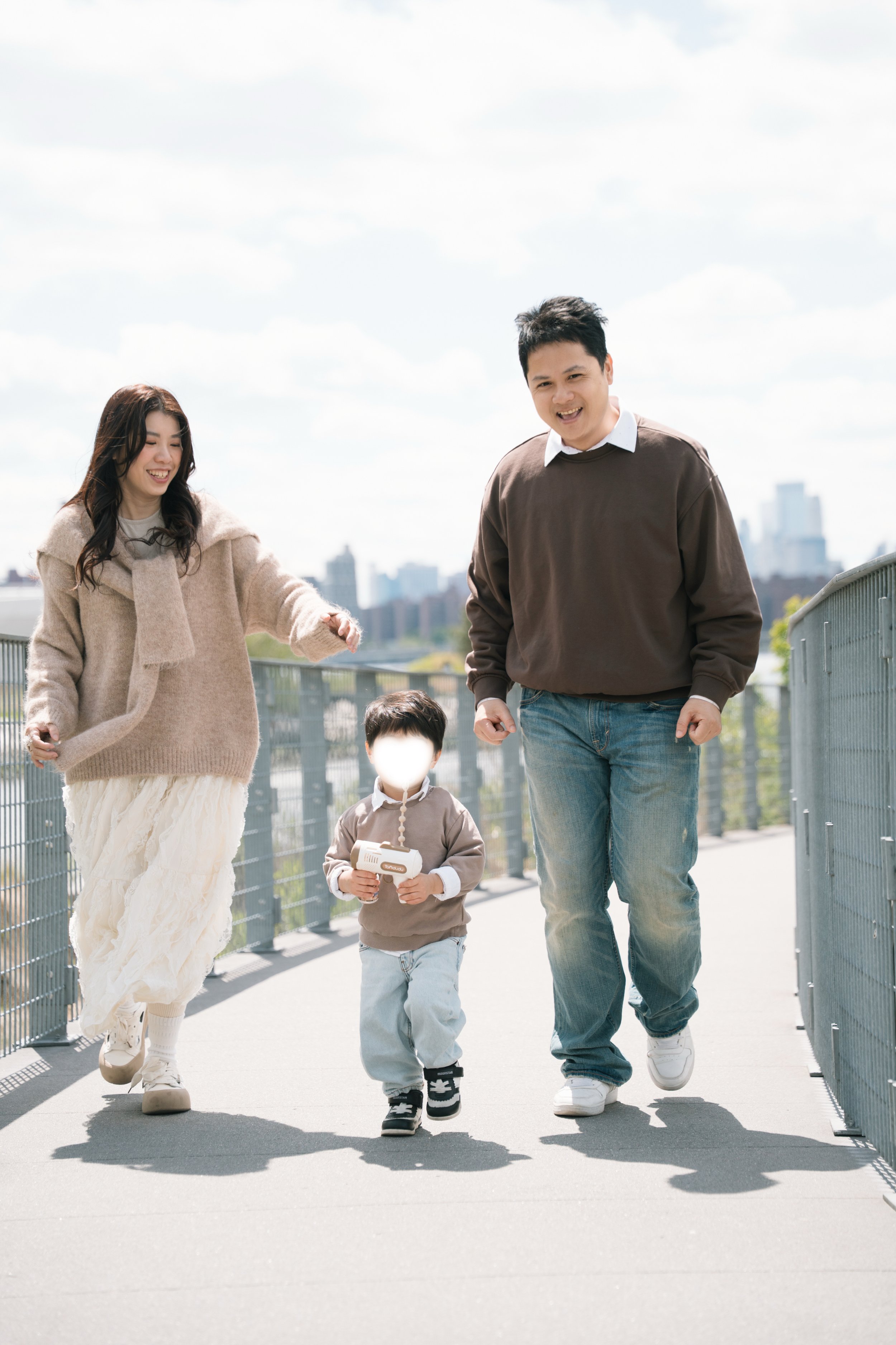 A family of four walking outdoors on a bright day, with a city skyline in the background. The mother is smiling and holding the hand of a young boy, who is holding a toy. The father is smiling and walking alongside them.
