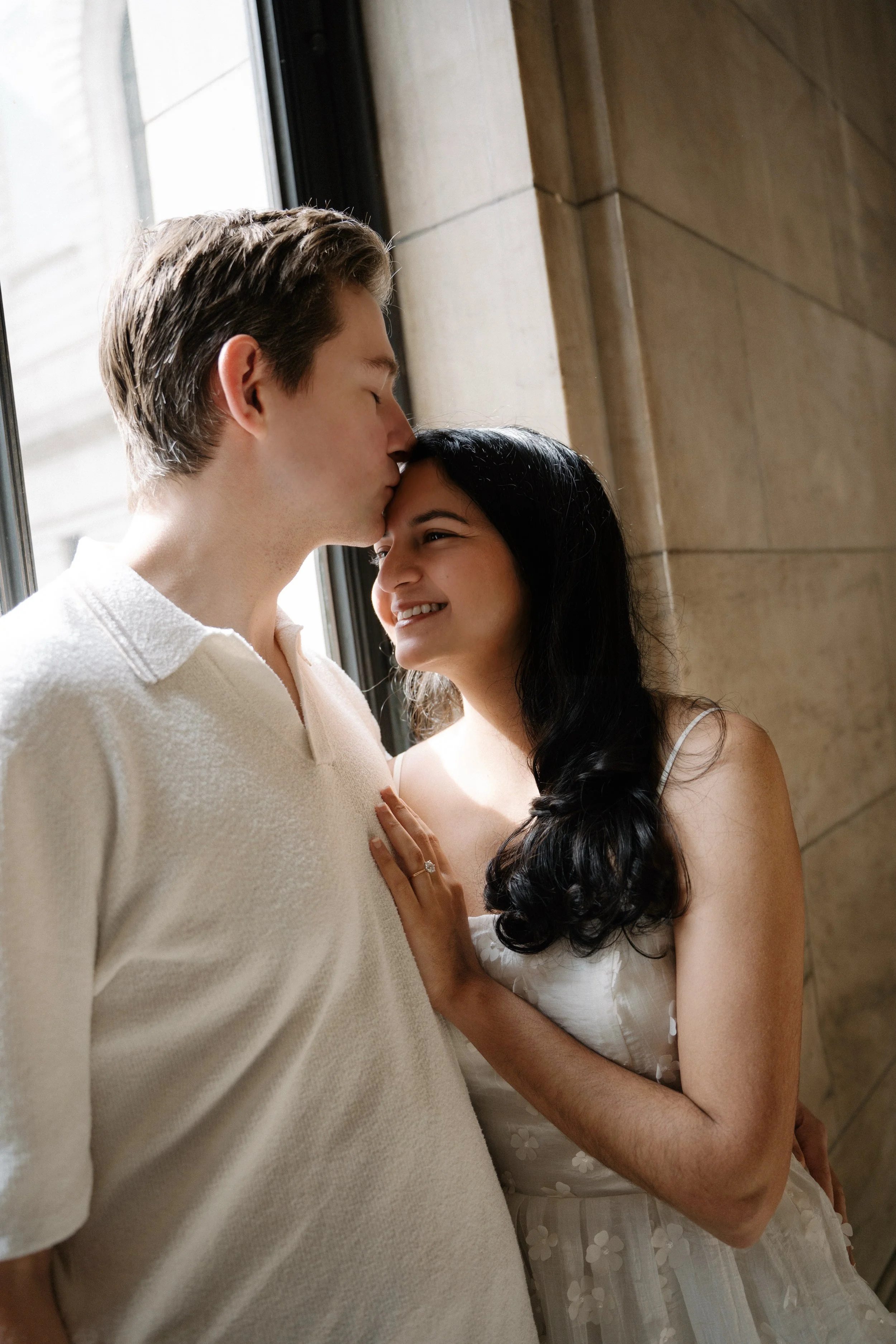 A couple standing close together indoors, with the man kissing the woman's forehead and the woman smiling with her hand on his chest.