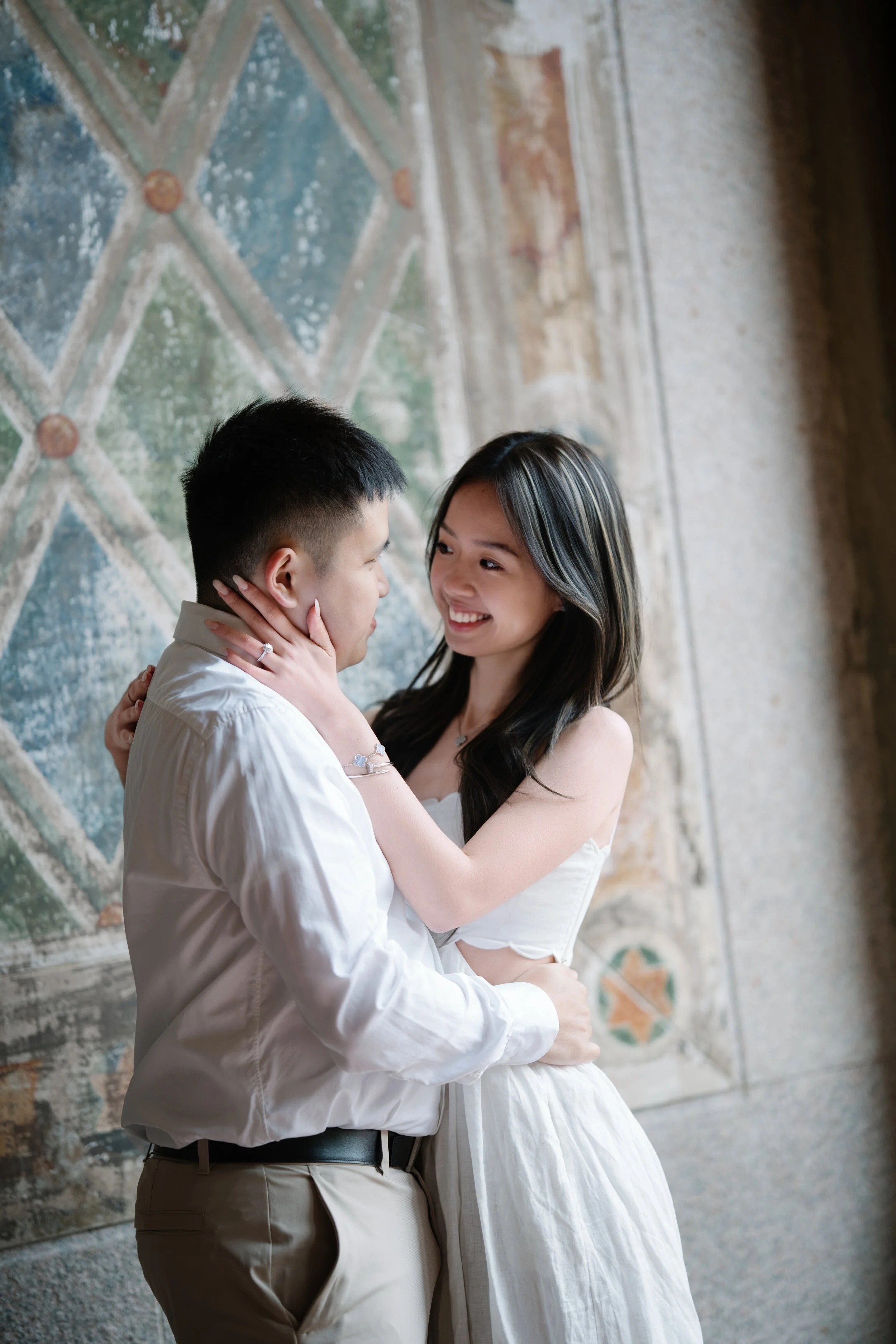 A couple standing close together, smiling and looking into each other's eyes, indoors with a decorative wall in the background, dressed in white attire.