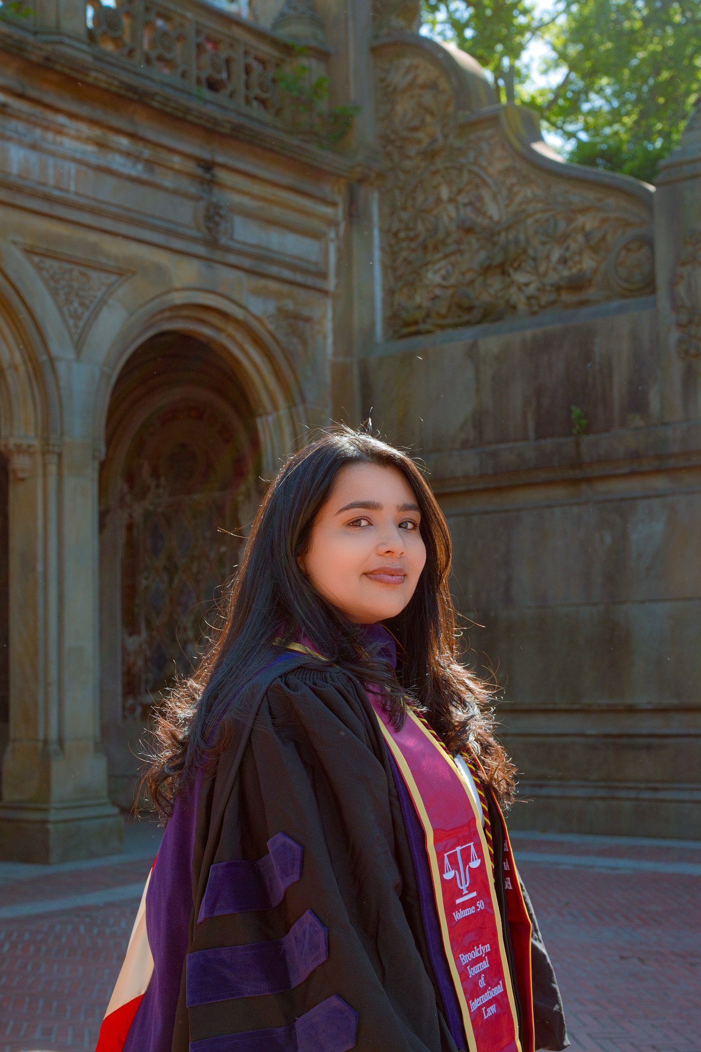 A young woman in graduation attire, including a stole with red and purple accents, stands outdoors in front of an ornate stone building with lush green trees overhead.