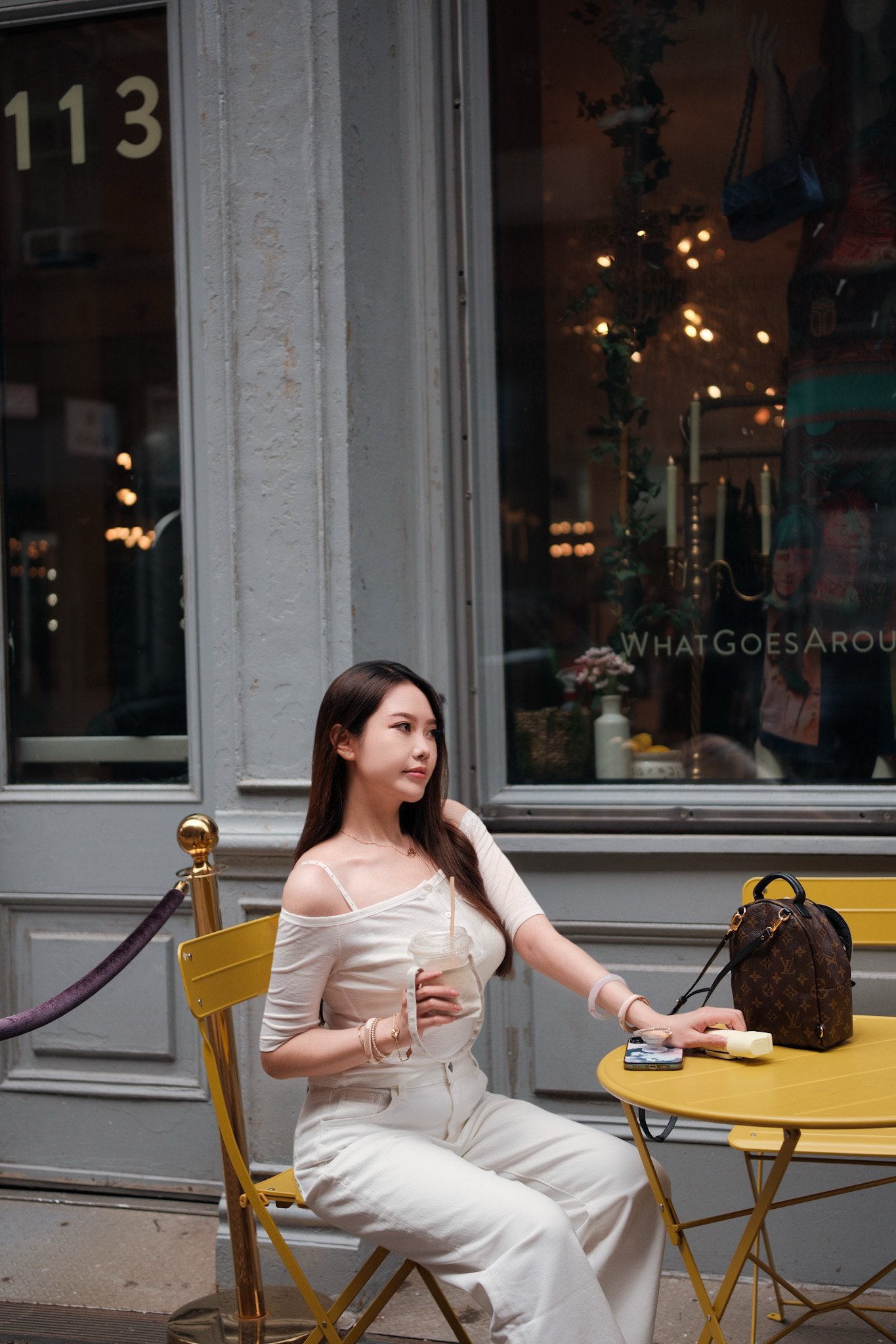 A young woman with long dark hair sitting outside at a yellow table at a cafe, holding a drink, with belongings including a Louis Vuitton bag and a phone on the table, and a window behind her reflecting lights.