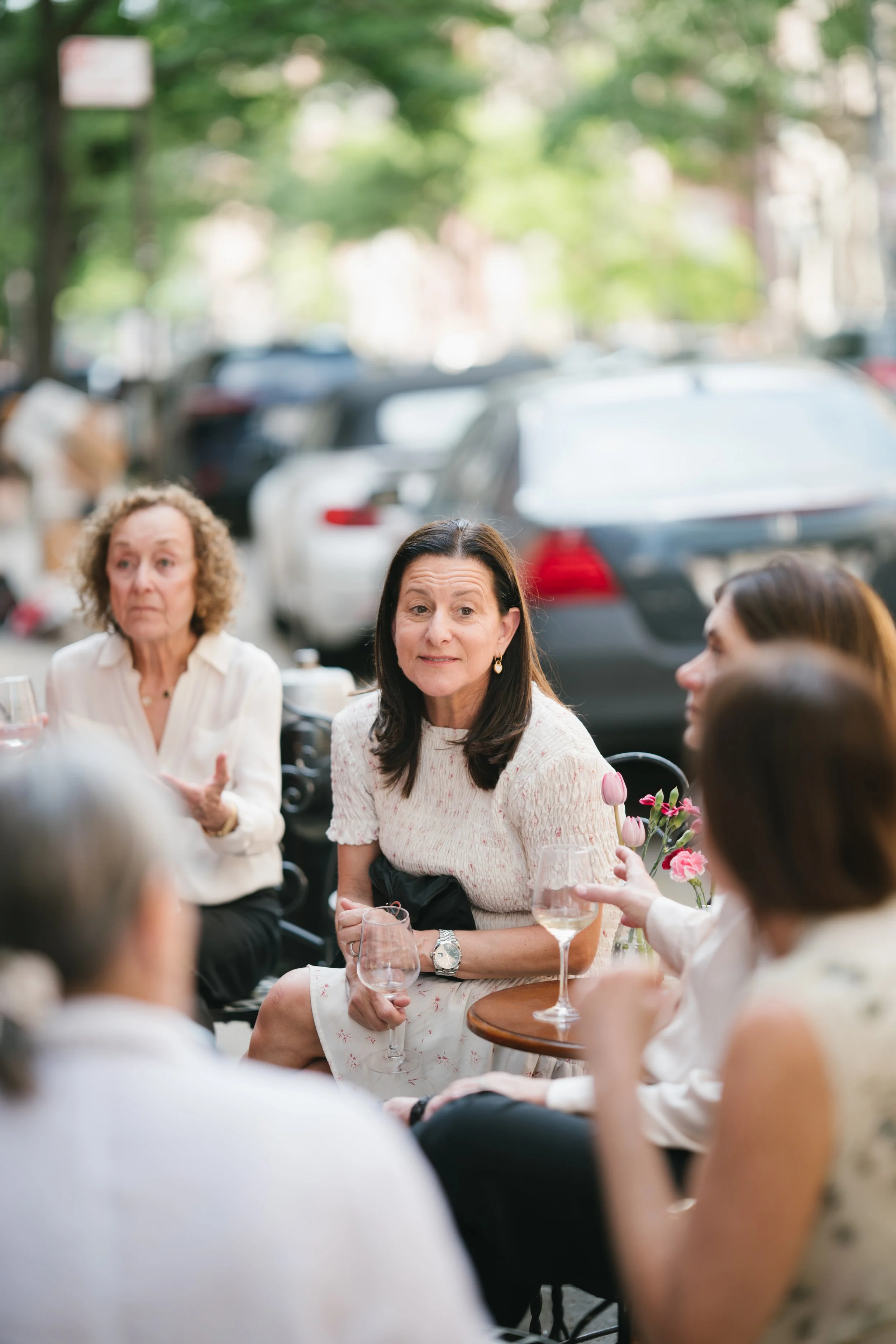 A group of women sitting outdoors at a social gathering, with some holding wine glasses and a woman in the center wearing a light pink dress and earrings, midst conversation. The background features parked cars and trees.