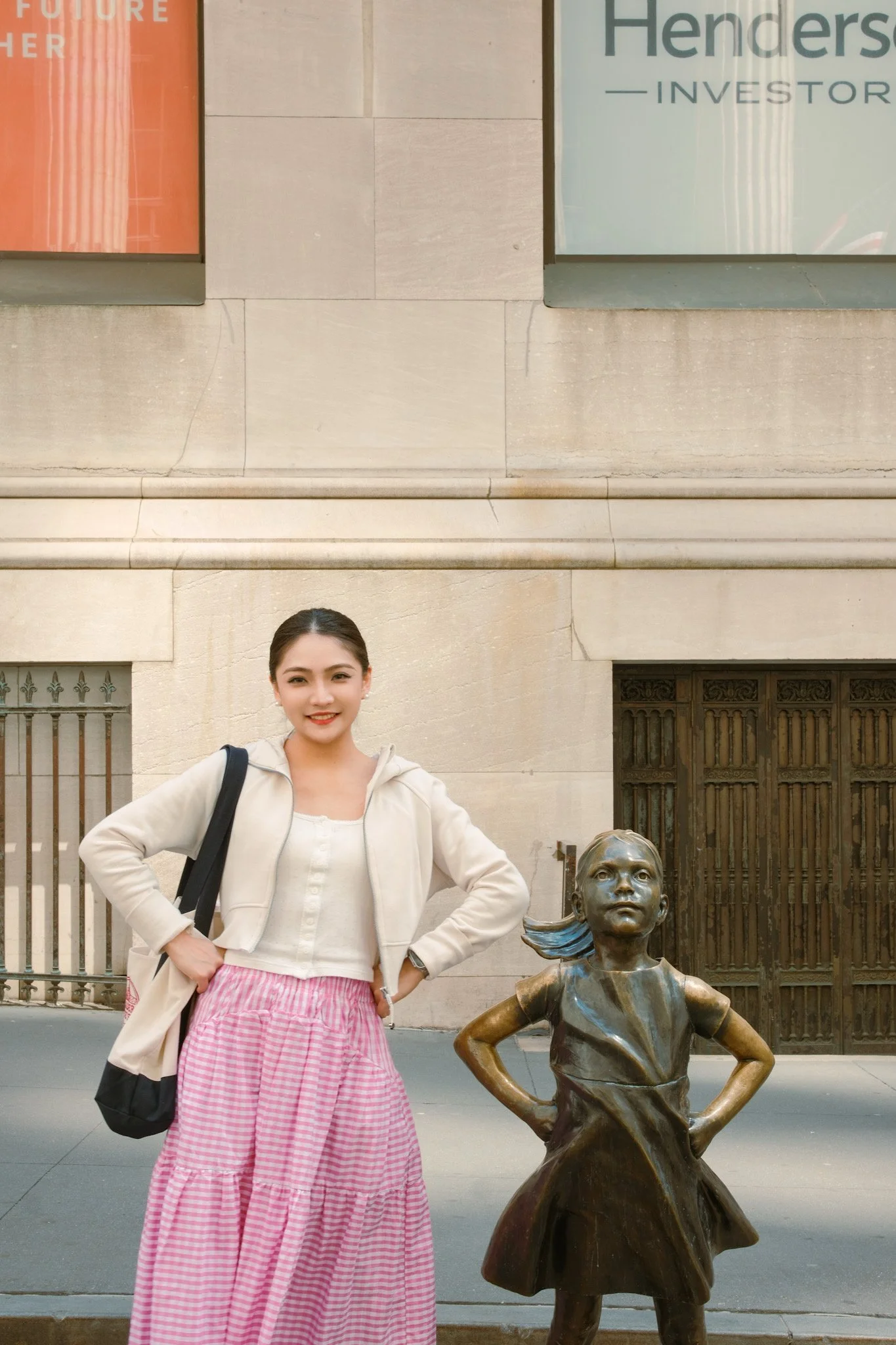 A young woman standing next to a statue of a girl with her hands on her hips in an urban setting.