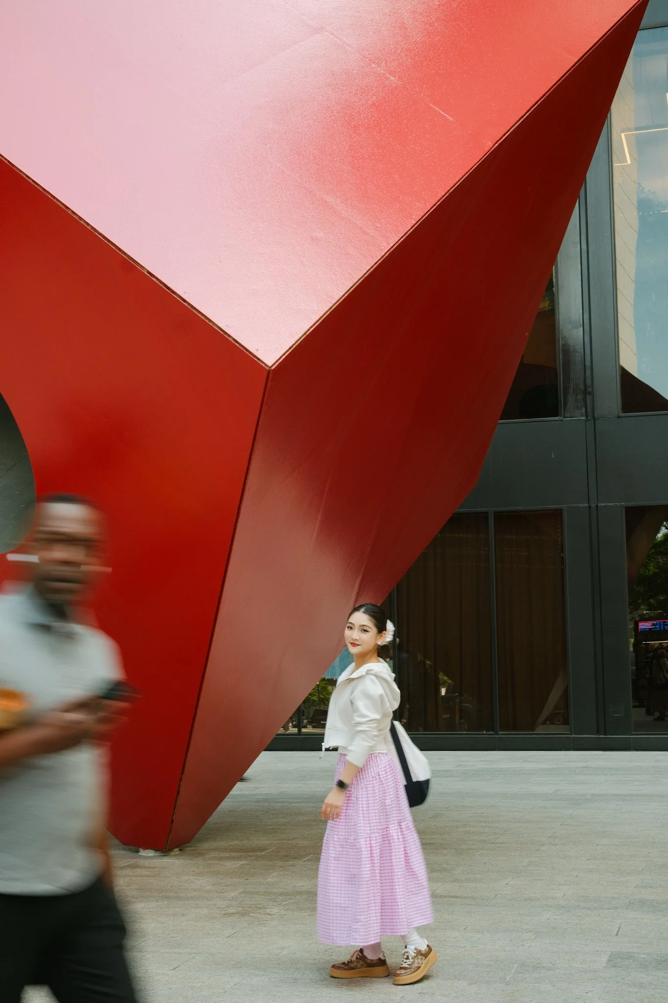 A young woman wearing a white jacket, pink and white checkered skirt, and brown sneakers stands near a red modern building, smiling at the camera. blurred pedestrians are in motion passing by.