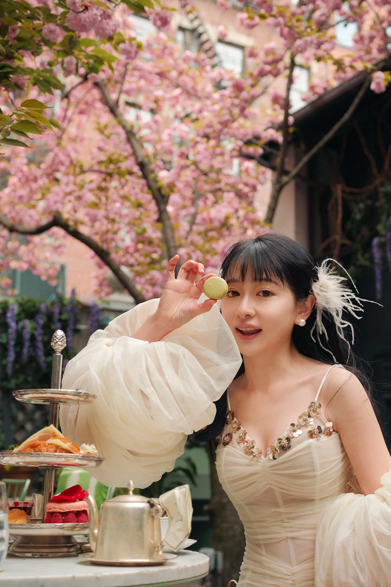 Young woman in a white dress with floral embellishments celebrating at an outdoor tea party, holding a green macaron. Pink flowering trees and a tiered tray with desserts are in the background.