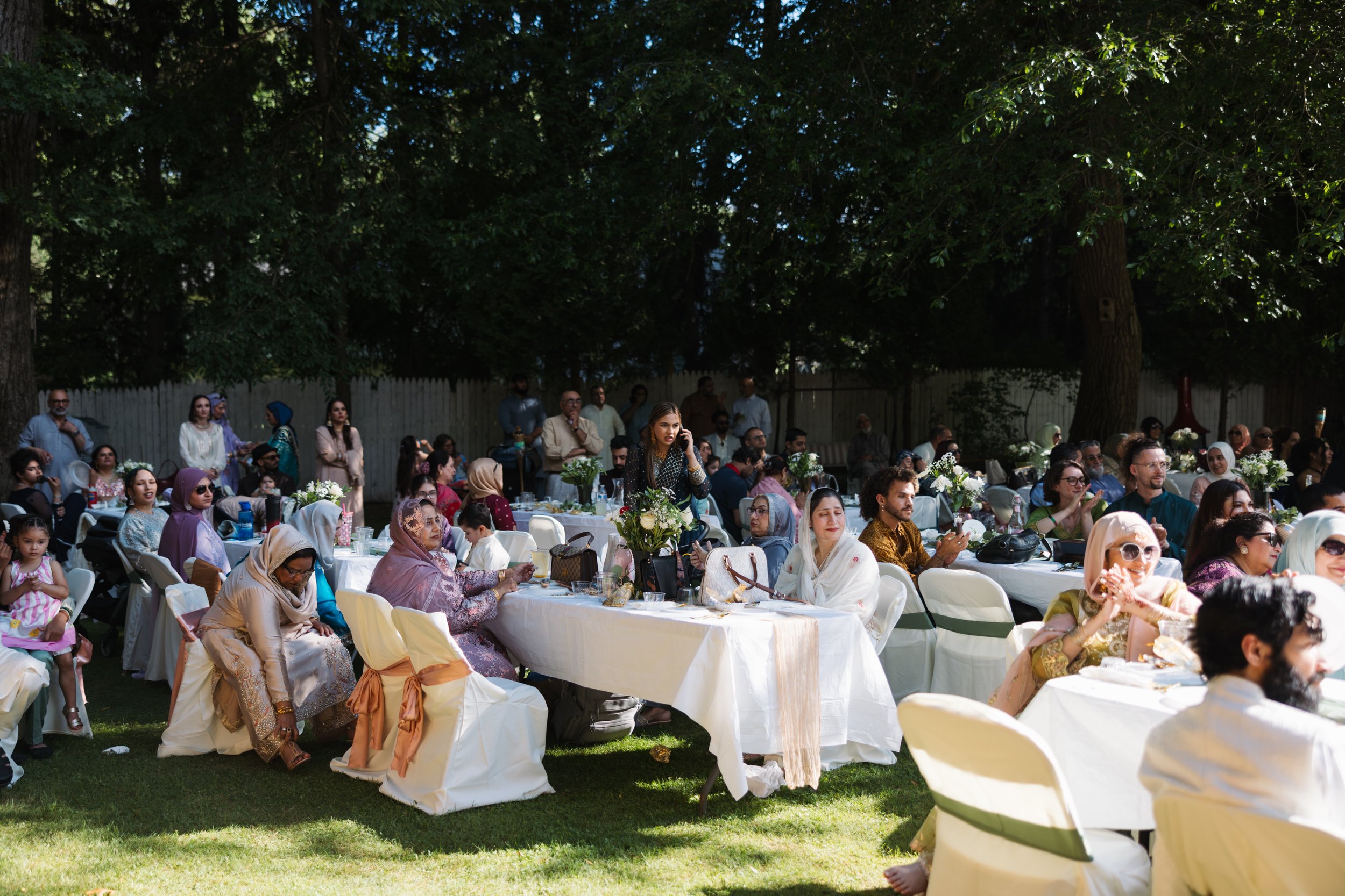 People seated at white tables outdoors during a gathering or celebration, with trees in the background.
