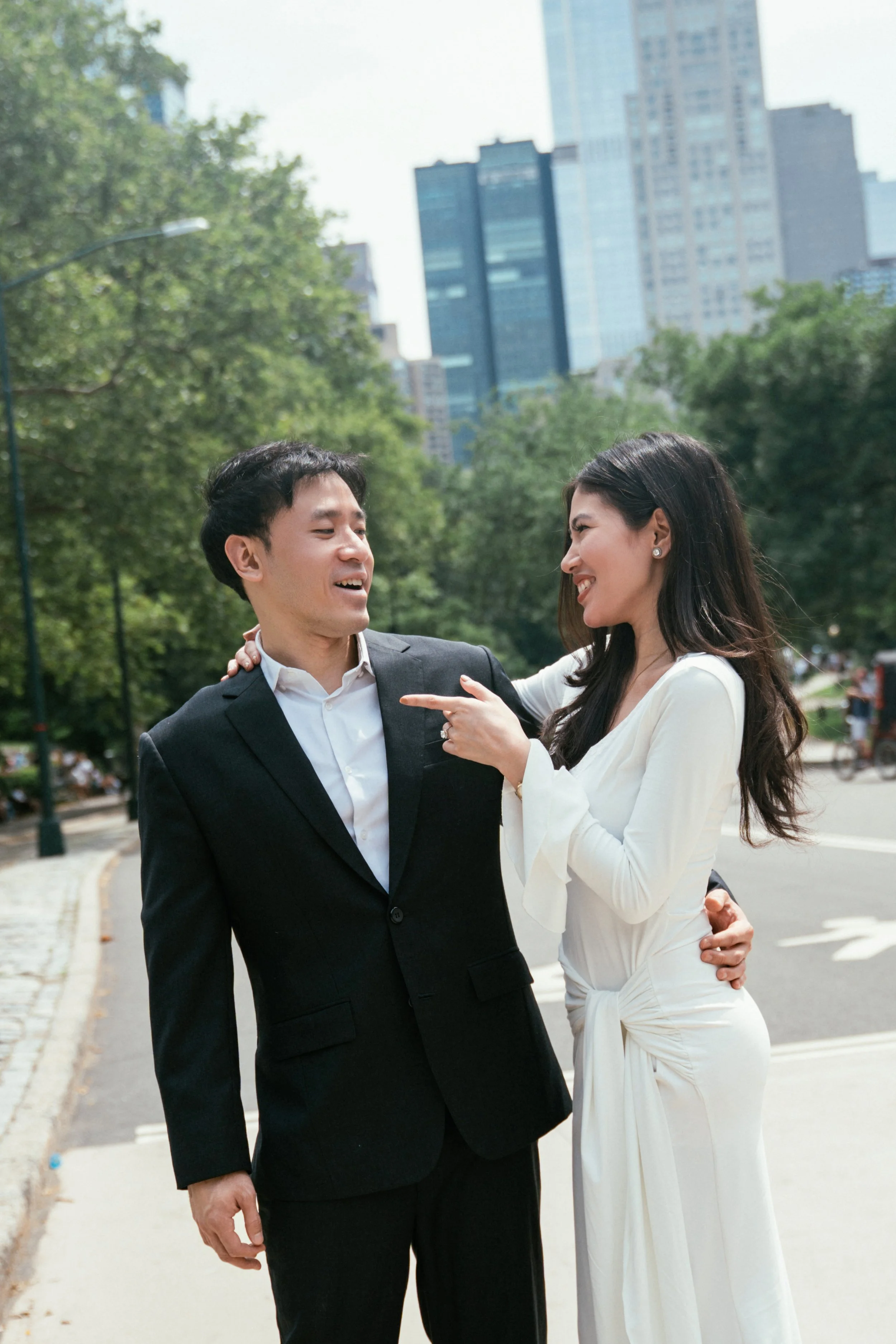 A man and woman dressed in formal attire sharing a joyful moment outdoors in a city park, with trees and skyscrapers in the background.