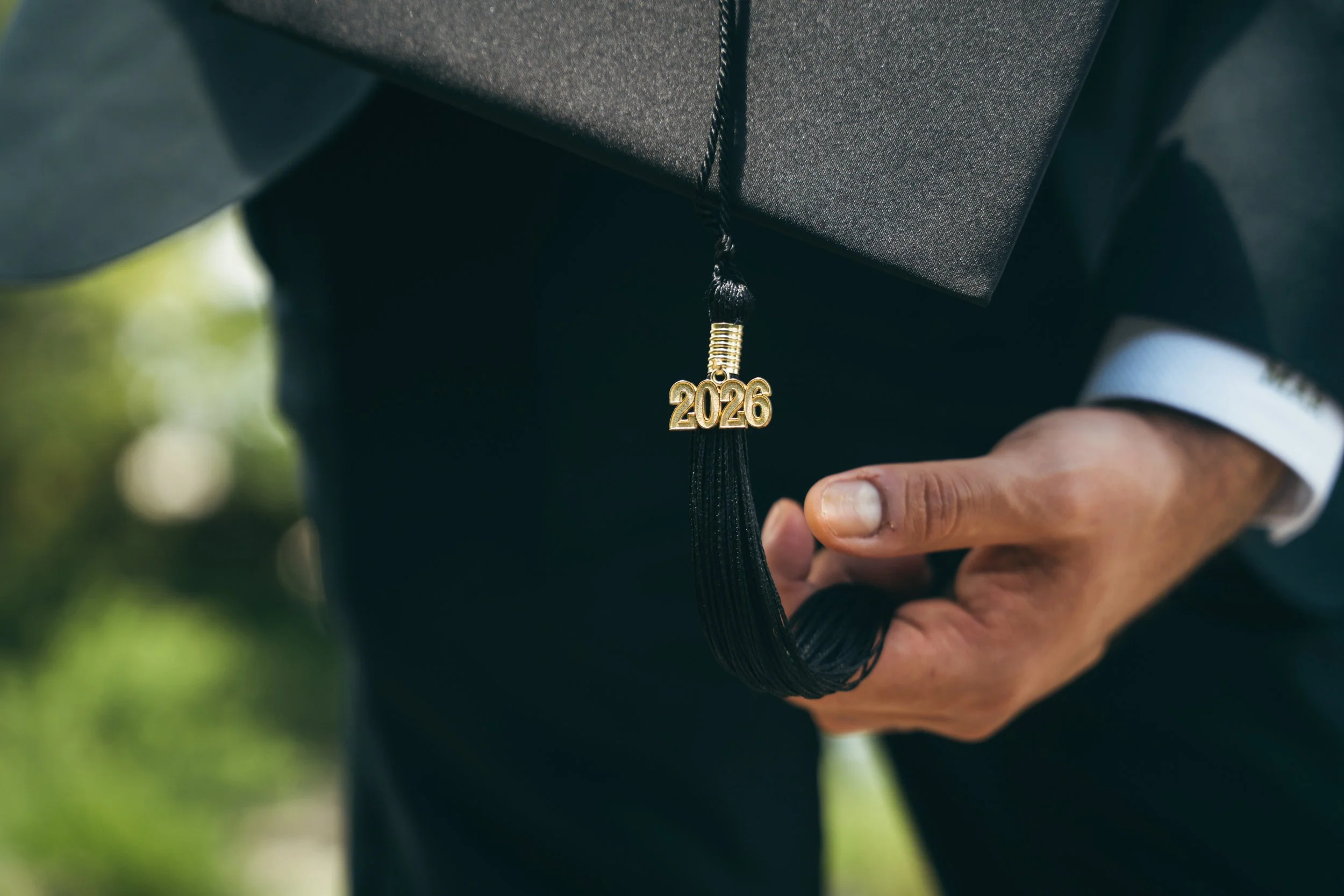 Close-up of a graduation cap with a black tassel and a gold '2026' charm. A person in formal attire is holding the cap, with a blurred outdoor background.