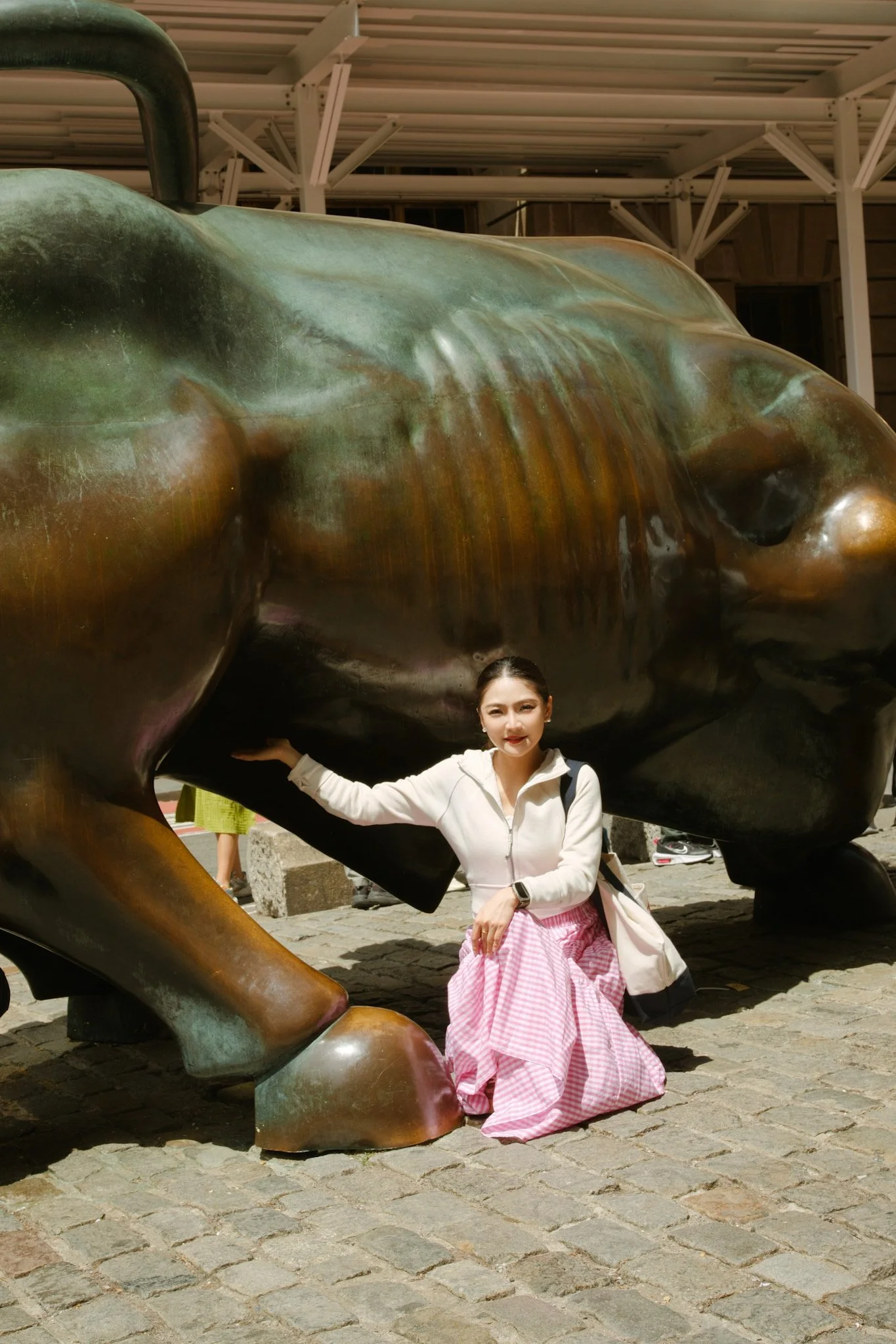 A woman is posing and smiling while crouching under a large bronze sculpture of a reclining figure with thick limbs. She is wearing a white jacket, pink and white chequered skirt, and has her hair tied back. The sculpture is outdoors, on a cobbleston