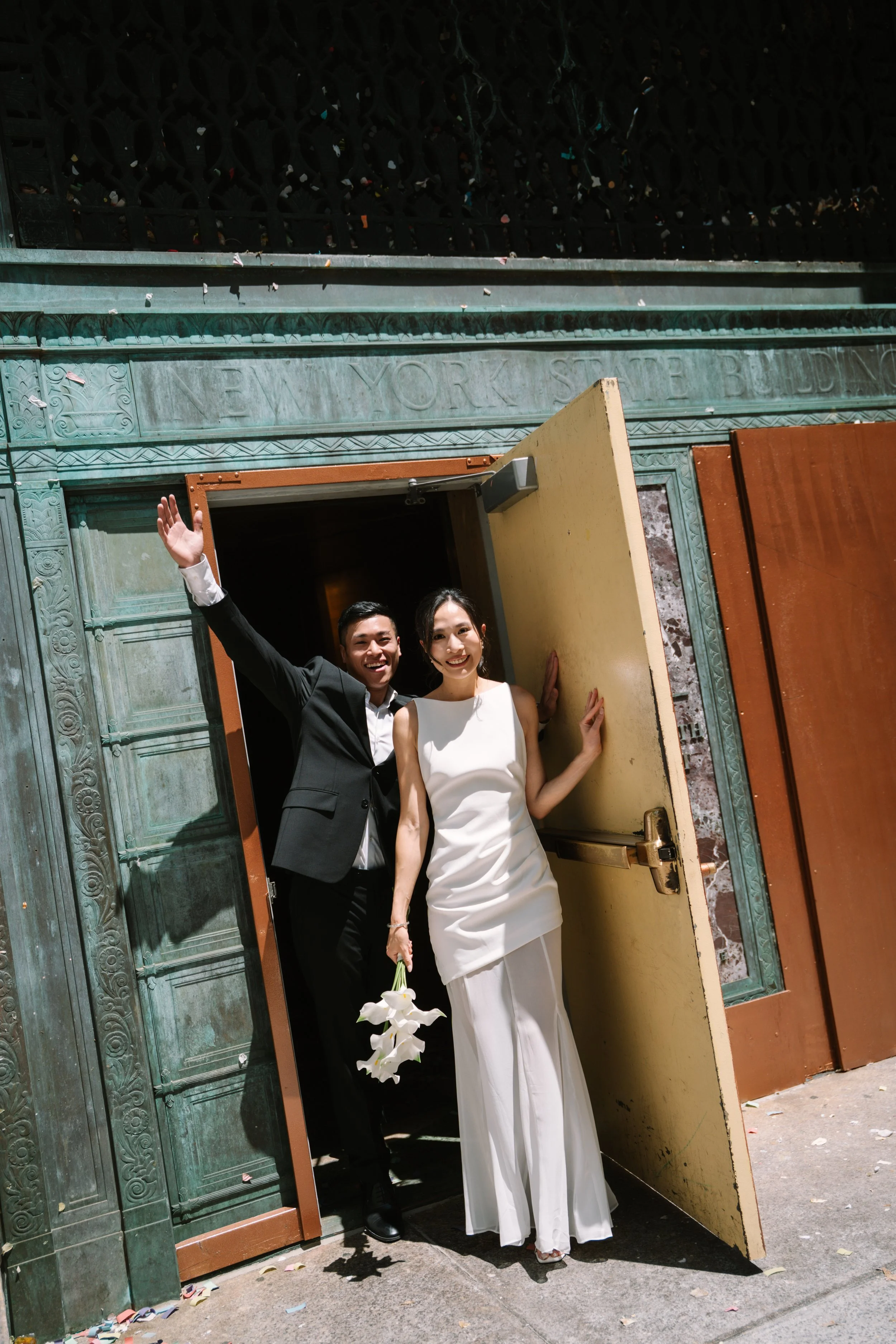 A happy couple, a man in a black tuxedo and a woman in a white wedding gown, standing in a doorway with a yellow door and green decorative wall, waving and holding a bouquet of white flowers.