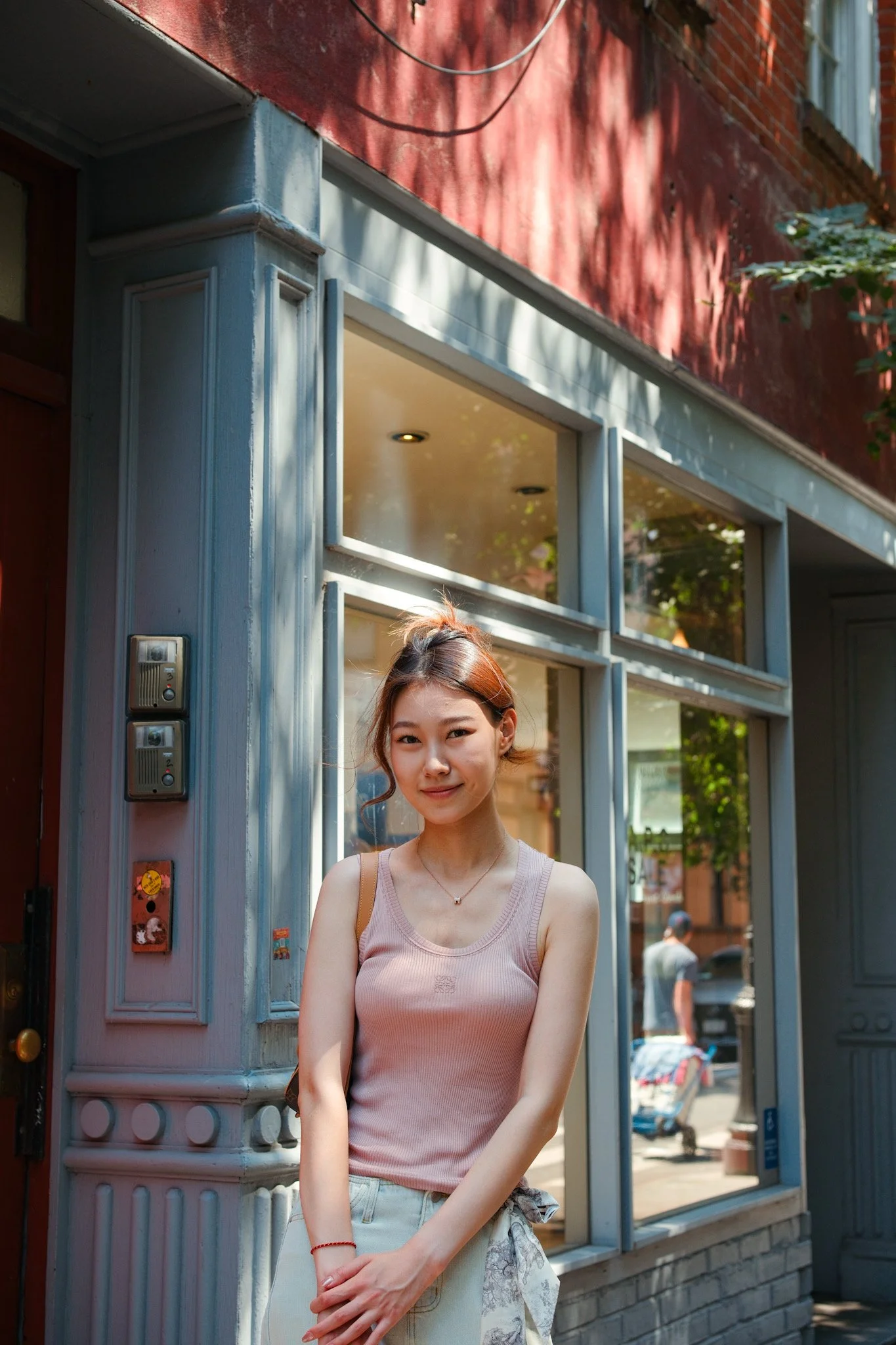 A young woman with light skin and brown hair tied in a casual bun, wearing a pink sleeveless top and gray skirt, standing outside on a city street near a building with red brick and blue-gray trim, smiling at the camera.