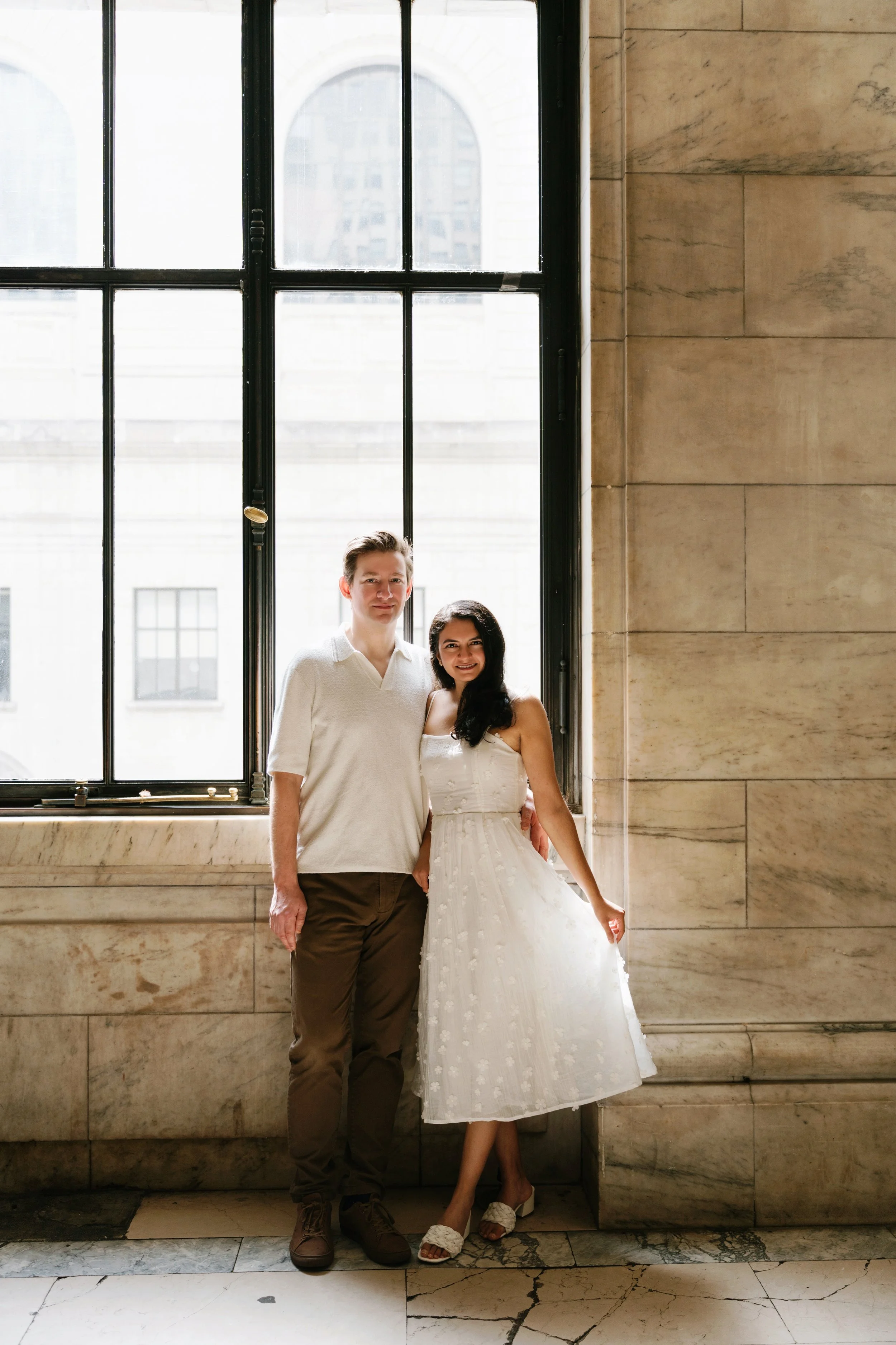 A man and woman standing together near a large window in a marble building, with the woman wearing a white dress and the man in casual brown pants and a white shirt.