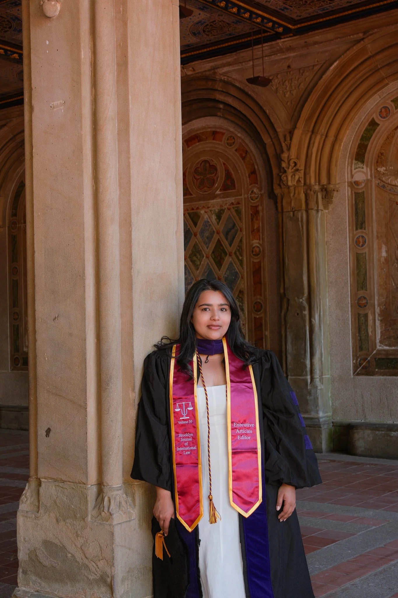A young woman in graduation attire, including a black gown and a maroon, purple, and gold sash, standing next to a stone pillar in a historic building with ornate architecture and colorful decorative murals on the walls.