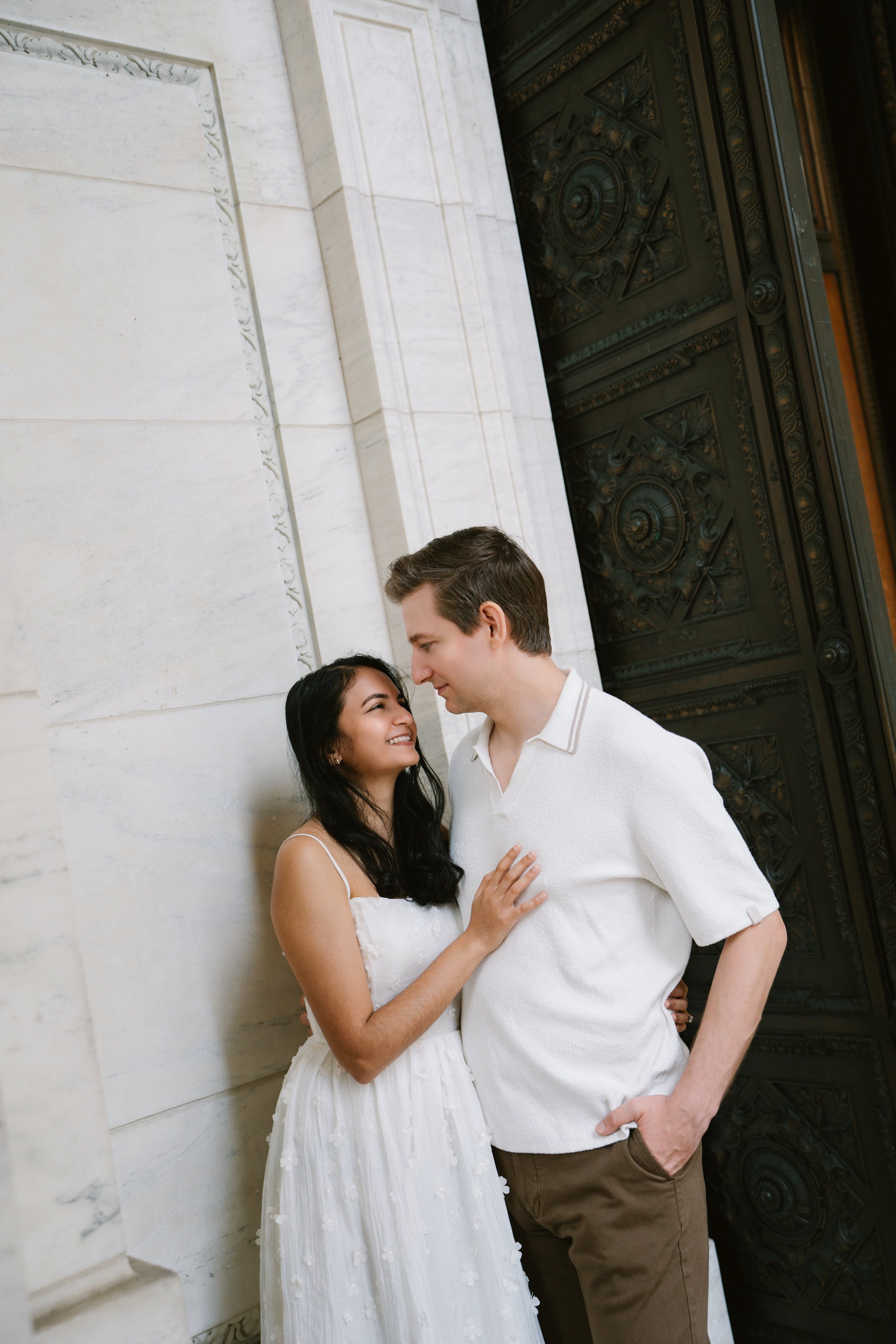 A young couple is standing close to each other, smiling and looking into each other's eyes, in front of a large, ornate black gate and a cream-colored marble wall.