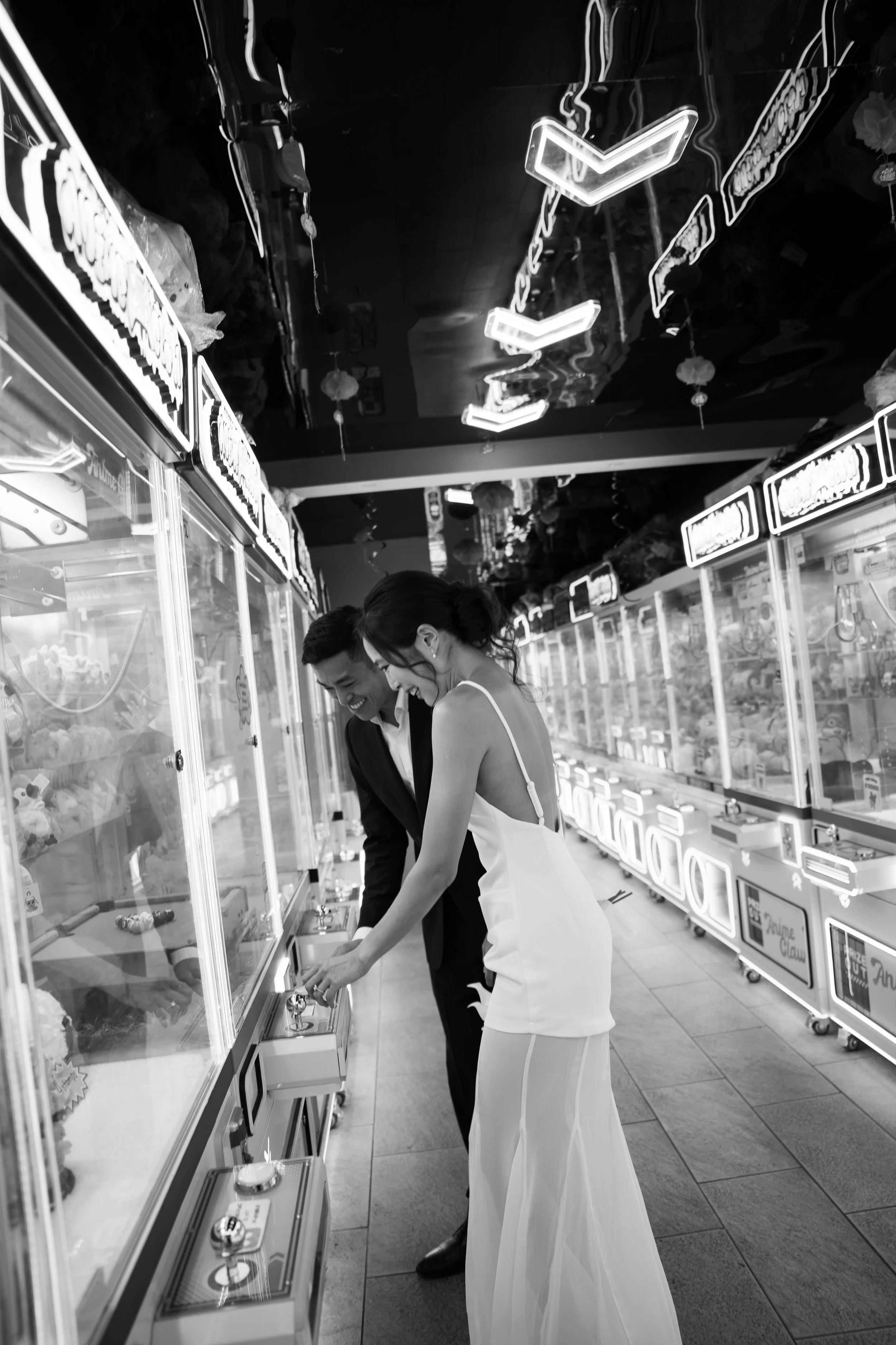 A man and woman dressed formally selecting jewelry from display cases in an arcade or mall with neon sign signs overhead.