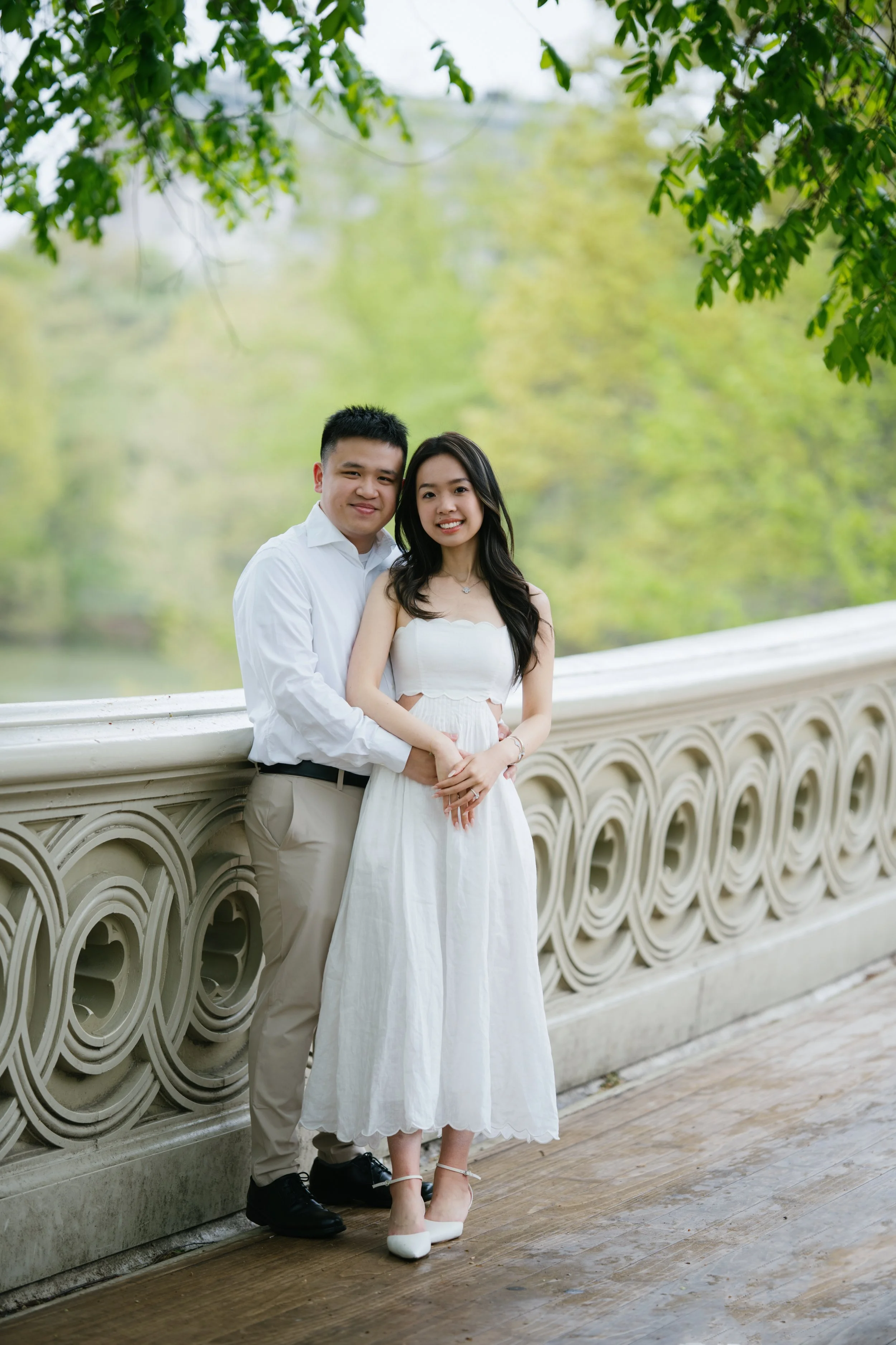 A couple standing close together outdoors on a bridge with decorative railing, surrounded by green trees and foliage, during daytime.