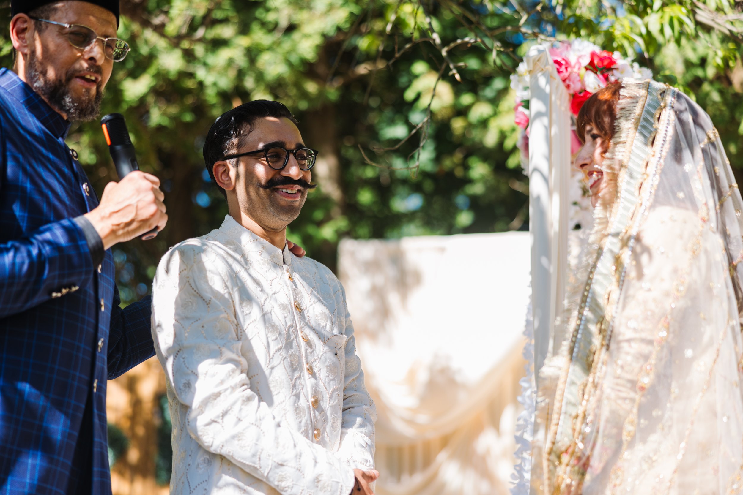 A groom and two men at an outdoor wedding ceremony, with the groom smiling and wearing traditional attire, and a woman in a richly embroidered dress facing him, under a canopy decorated with flowers.