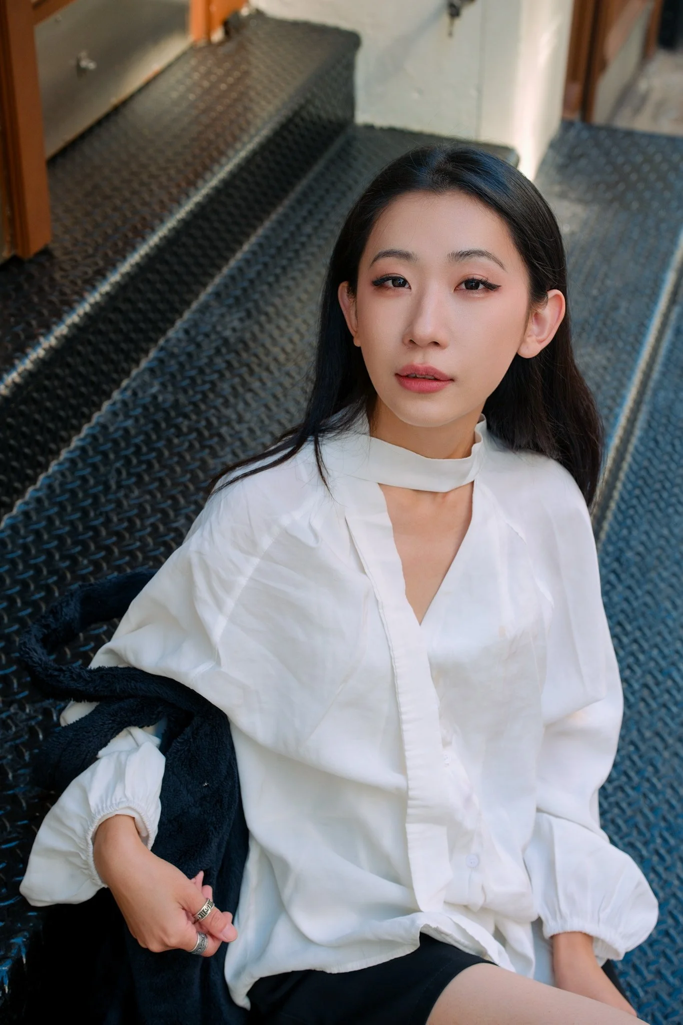 A young woman with long dark hair, wearing a white blouse, sitting outdoors on a black woven bench, with a black jacket draped over her lap, looking at the camera.