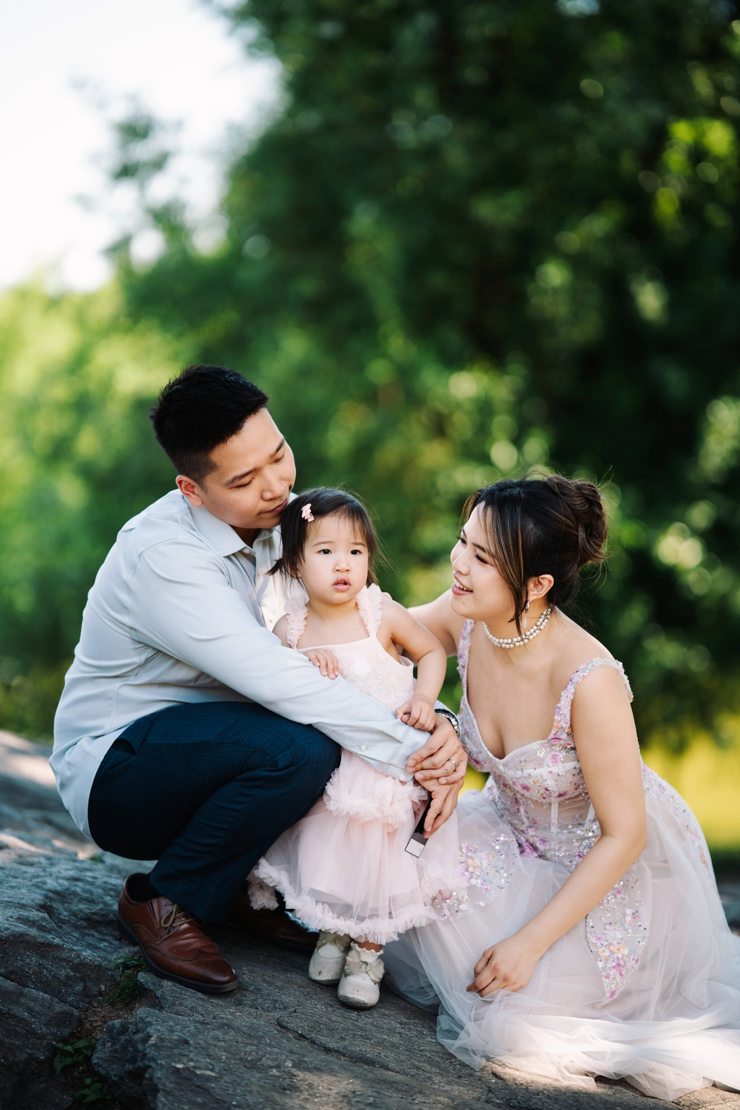 A family of three outdoors, with the father holding a young girl, the mother kneeling beside them, all looking at each other, with a background of green trees.