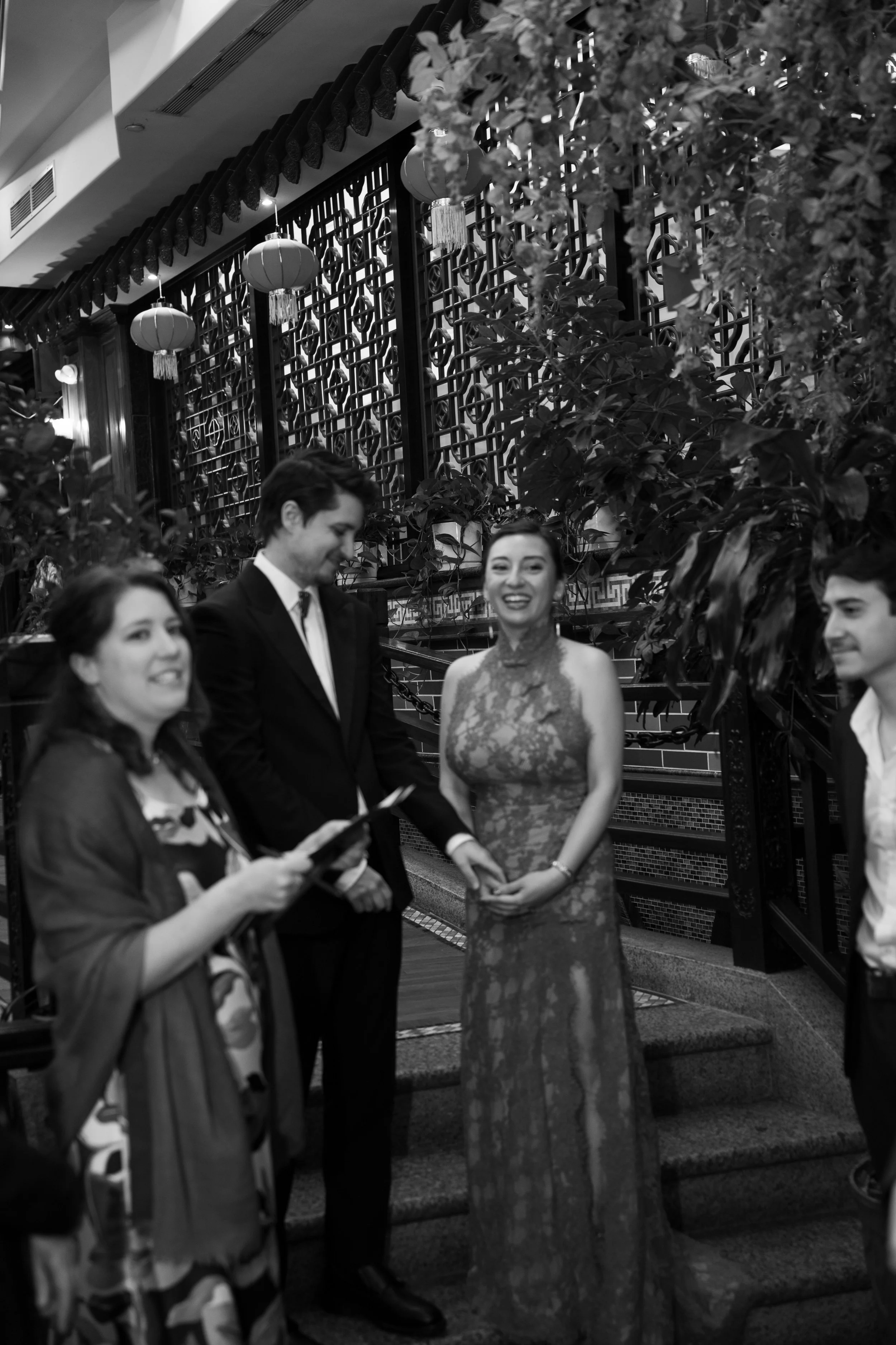 Group of young adults in formal attire standing on stairs in a decorated indoor setting, possibly at a celebration or event, with traditional Chinese lanterns and decorative elements in the background.