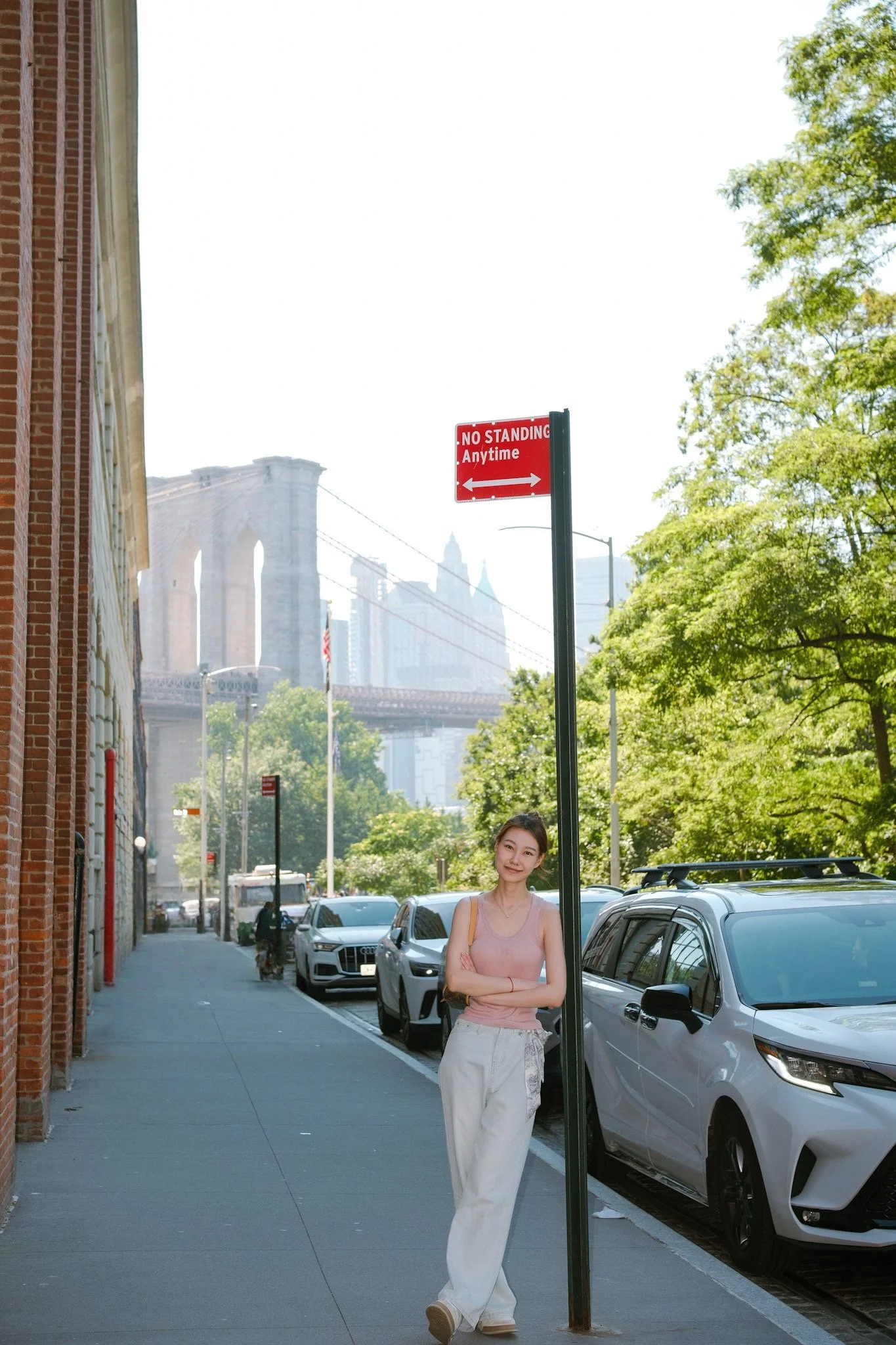 A young woman with a sleeveless pink top and white pants standing on a city sidewalk, leaning against a pole with a "No Standing Anytime" sign, with the Brooklyn Bridge and city buildings in the background