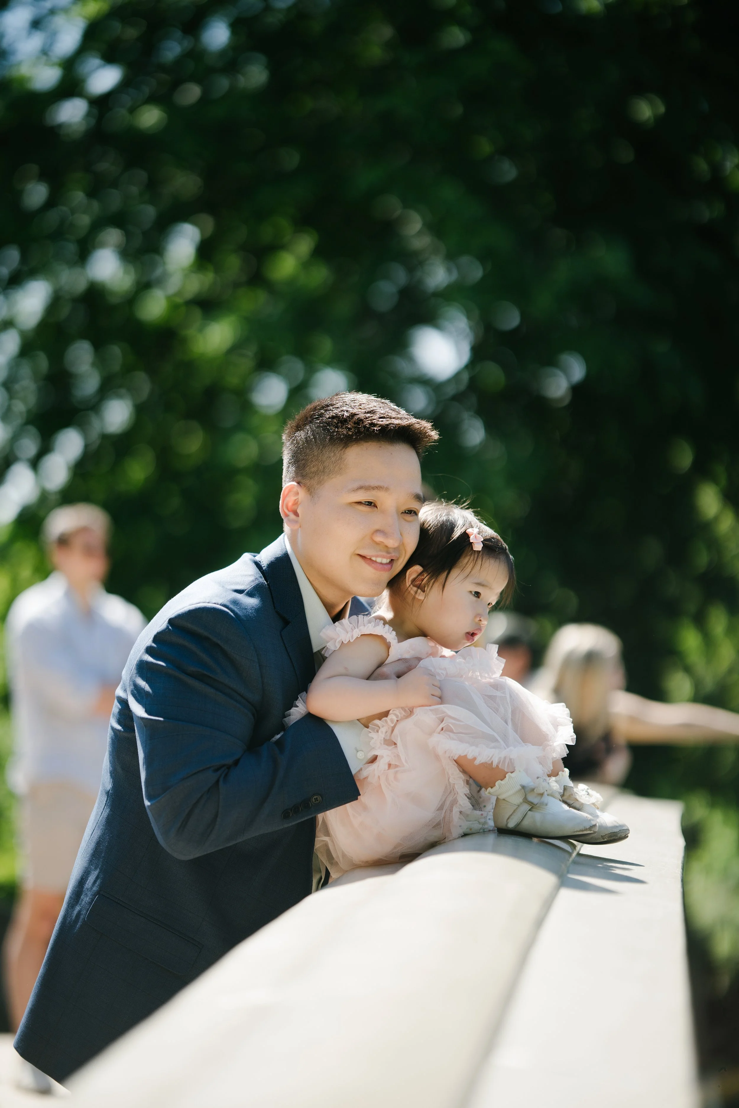 A man in a suit holding a young girl in a pink dress while crossing a bridge outdoors during daytime; people and trees are blurred in the background.