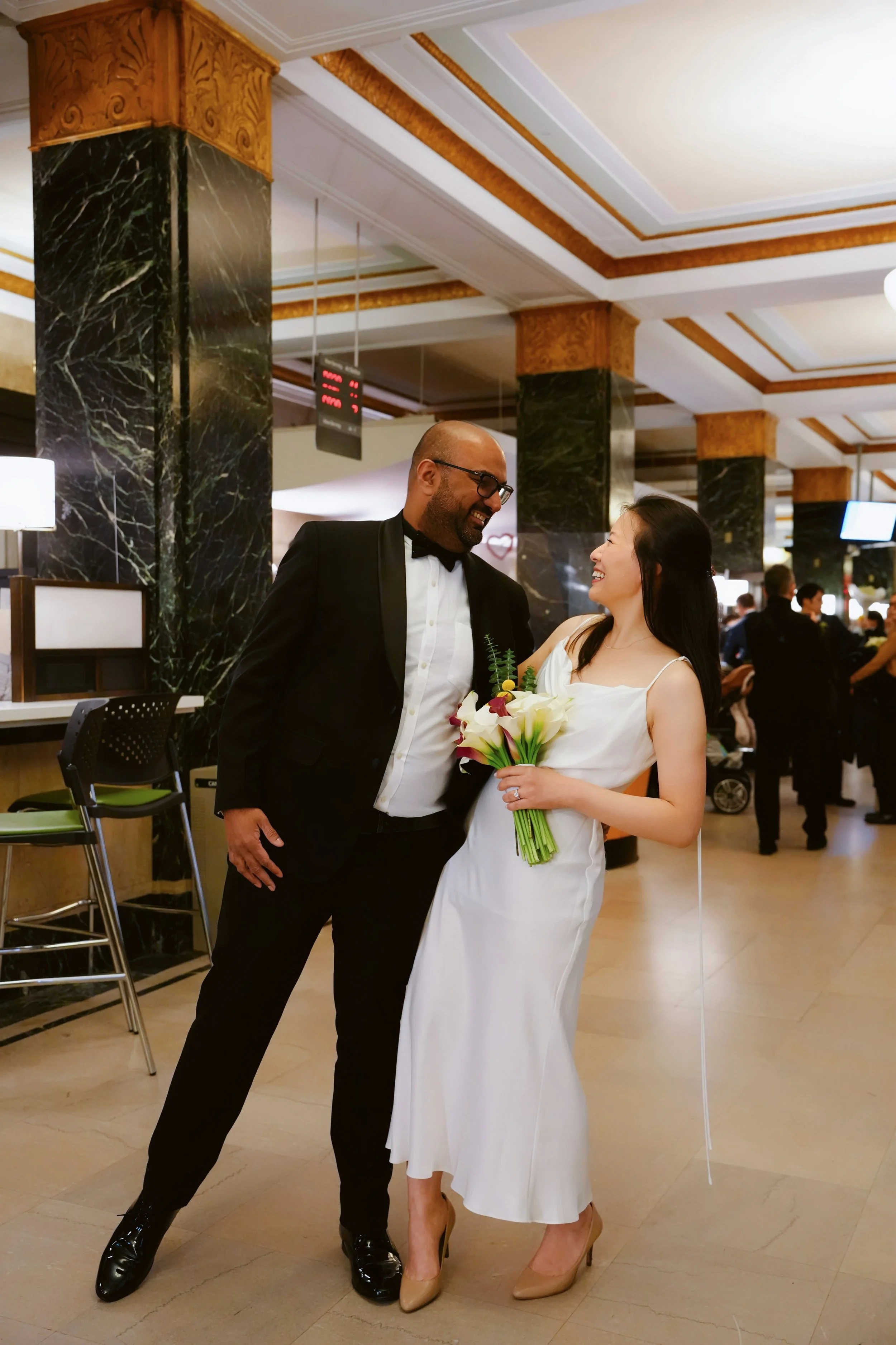 A man in a tuxedo and a woman in a white dress are smiling at each other in a decorated interior space, possibly a hotel or event venue, with other people in the background.