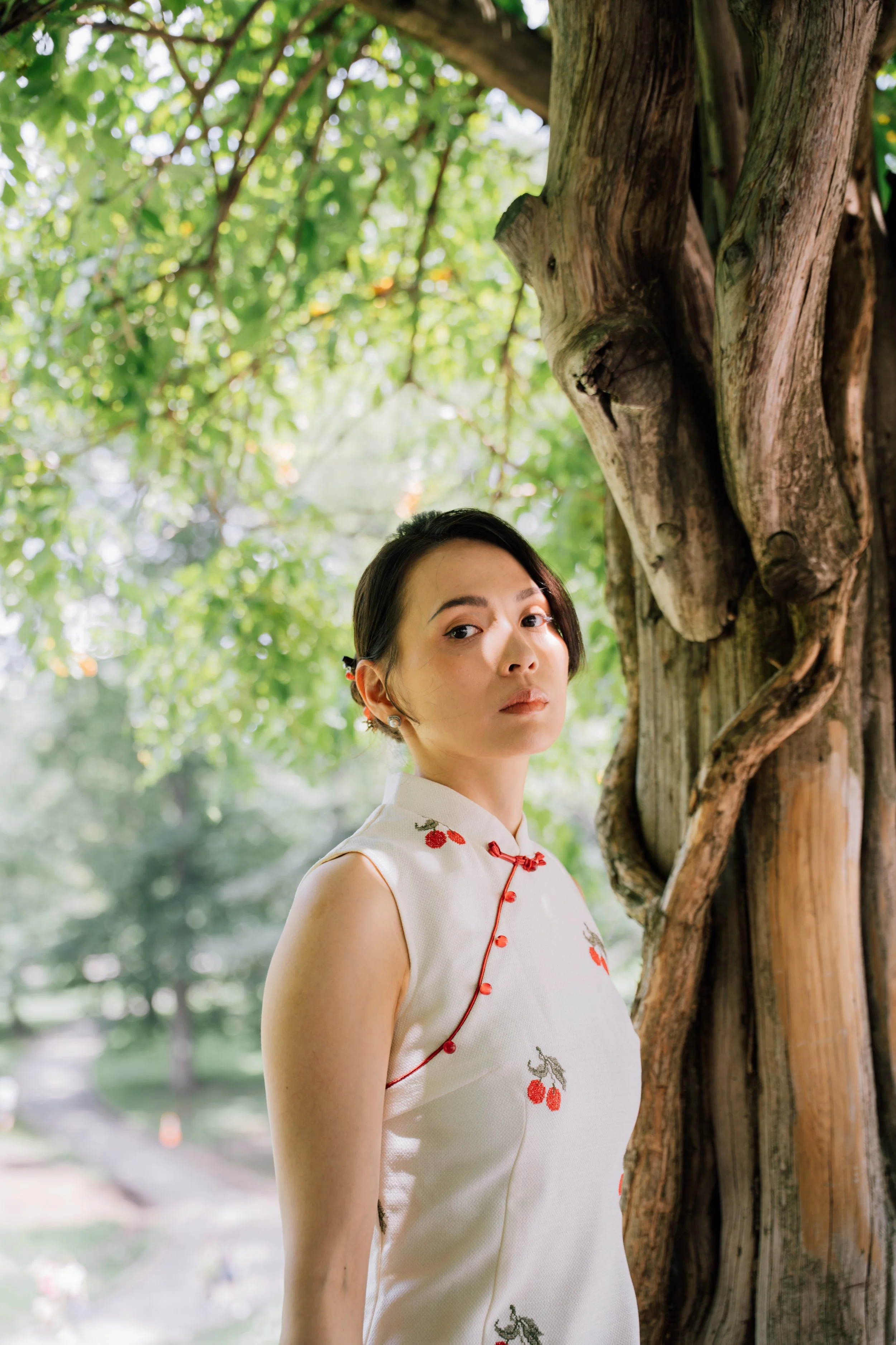A woman with short dark hair and earrings standing outdoors near a tree with a rough bark trunk. She wears a sleeveless white dress with red cherry embroidery and red piping, and gazes at the camera with a neutral expression. Green foliage and blurre