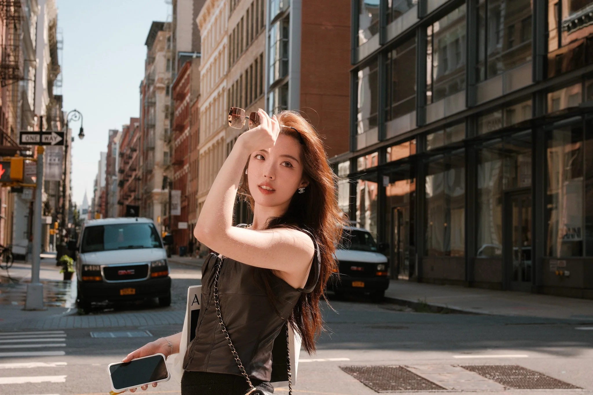 A young woman with long reddish-brown hair and fair skin is standing on a busy city street, holding sunglasses to her forehead with her right hand. She is wearing a black sleeveless top and carrying a white bag with black and white straps over her sh