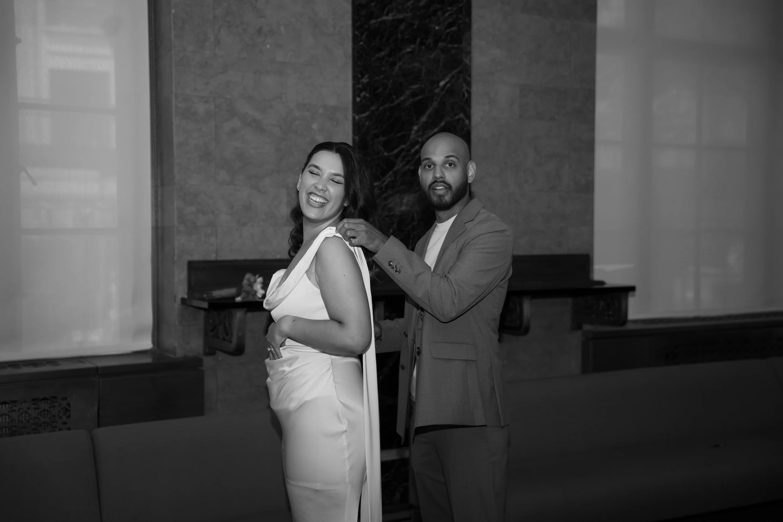 A woman in a white dress with a smiling expression and a man in a suit, both in an elegant indoor setting with marble walls, possibly at a formal event or celebration.