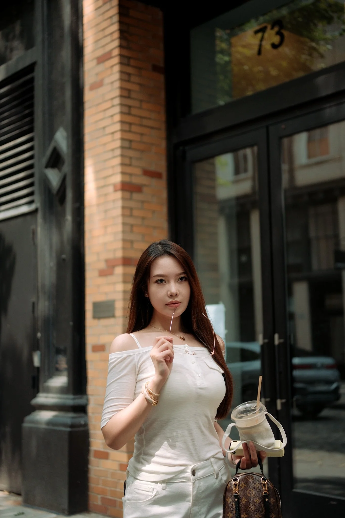 A young woman with brown hair wearing a white off-shoulder top and white pants is standing outside in front of a brick building with black window frames. She is holding a drink in one hand and a pair of sunglasses in the other, with a brown patterned