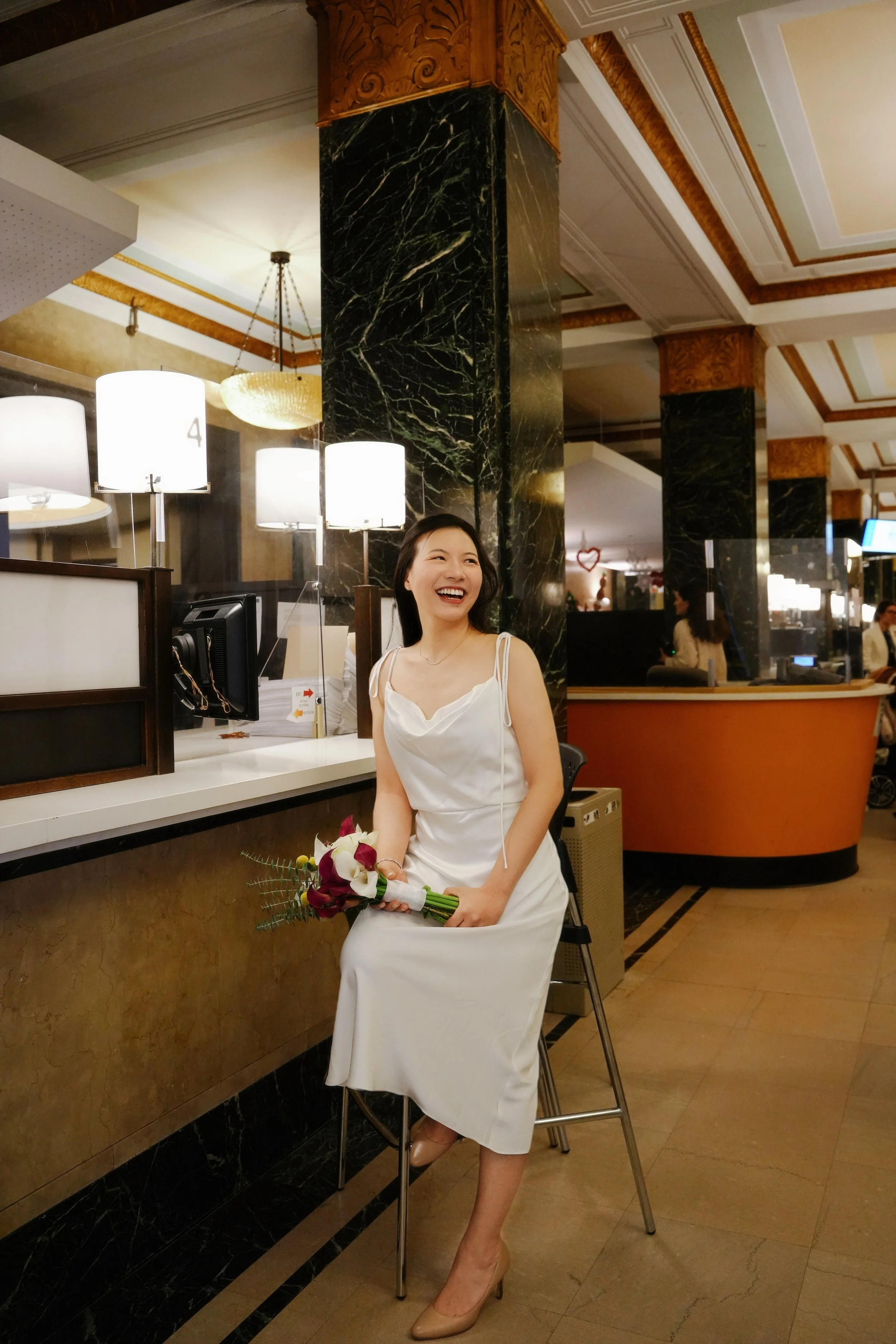 A woman in a white dress sitting on a chair in a hotel or reception area, holding a bouquet of flowers, smiling. The area has dark green marble columns, a reception desk, and warm lighting with ceiling fixtures. In the background, there are other peo