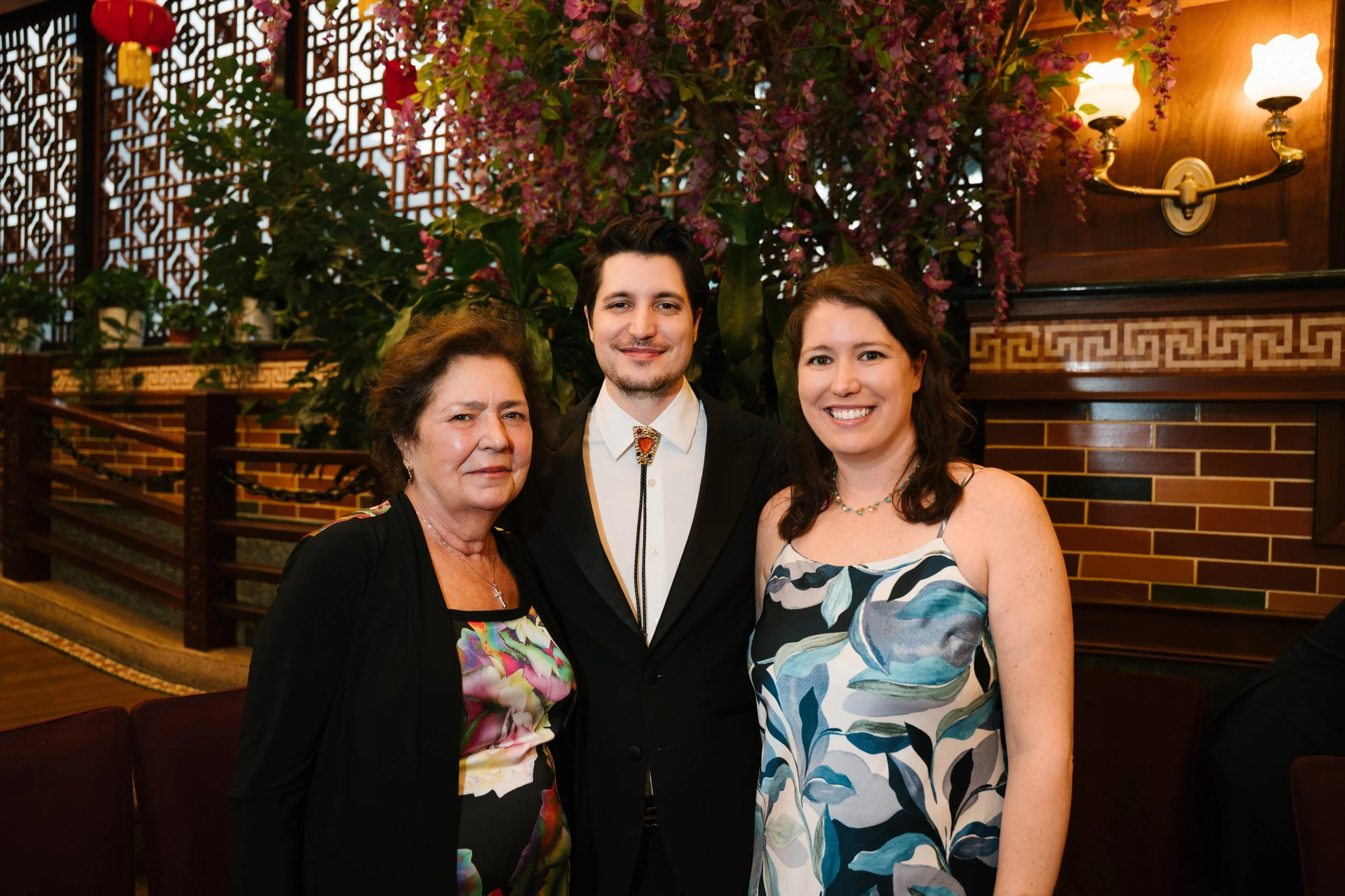 Three people standing close together in an indoor setting with decorative wooden lattice and large flowering plants behind them. They are smiling, with two women and one man in formal attire.
