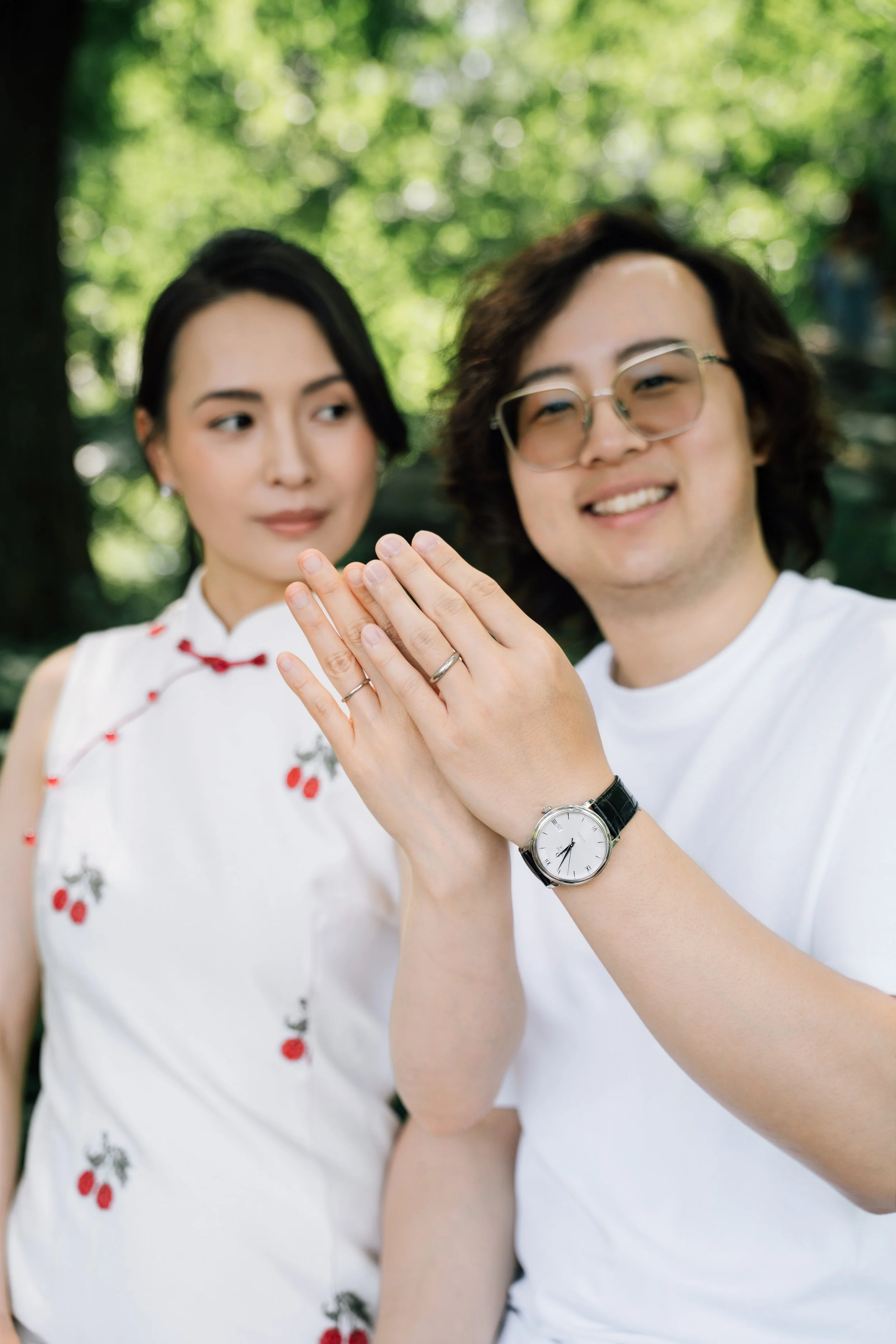 Two people showing off their wedding rings outdoors with green trees and sunlight in the background.