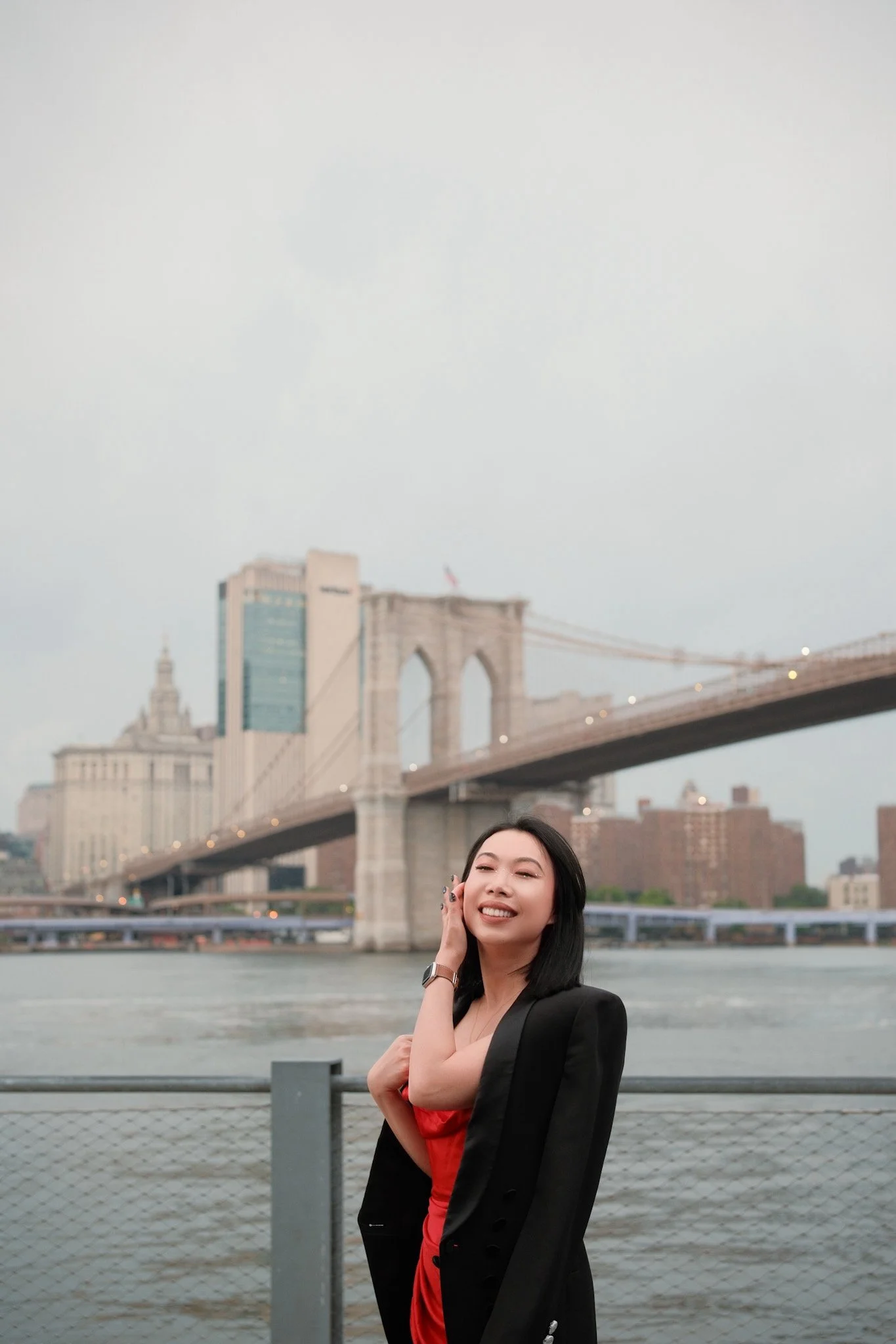 A woman standing by the water with the Brooklyn Bridge and Manhattan skyline in the background, smiling and posing with one hand near her face.