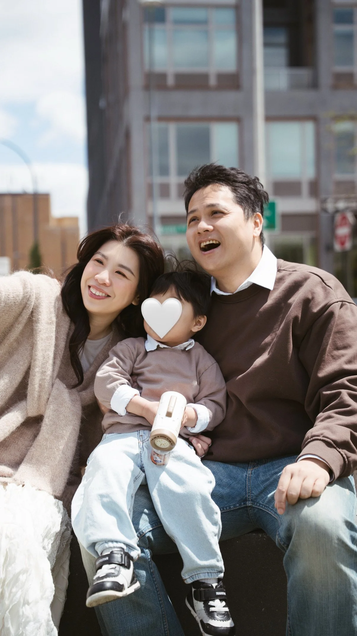 A happy family of three taking a selfie outdoors in an urban area with modern buildings in the background. The family includes a woman, a man, and a young boy with a heart shape over his face.