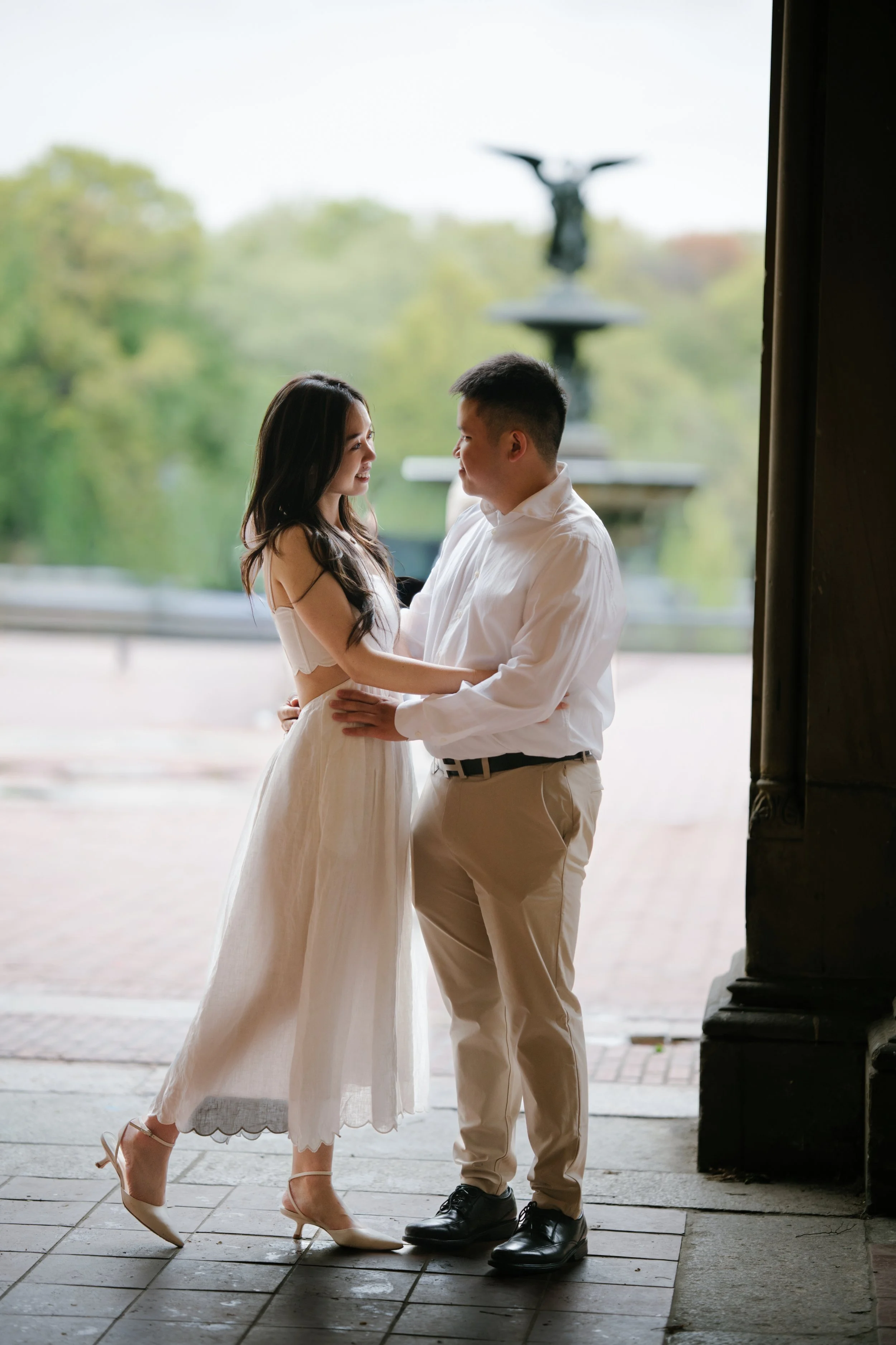 A couple dancing outdoors near a fountain, with trees in the background, during daytime.