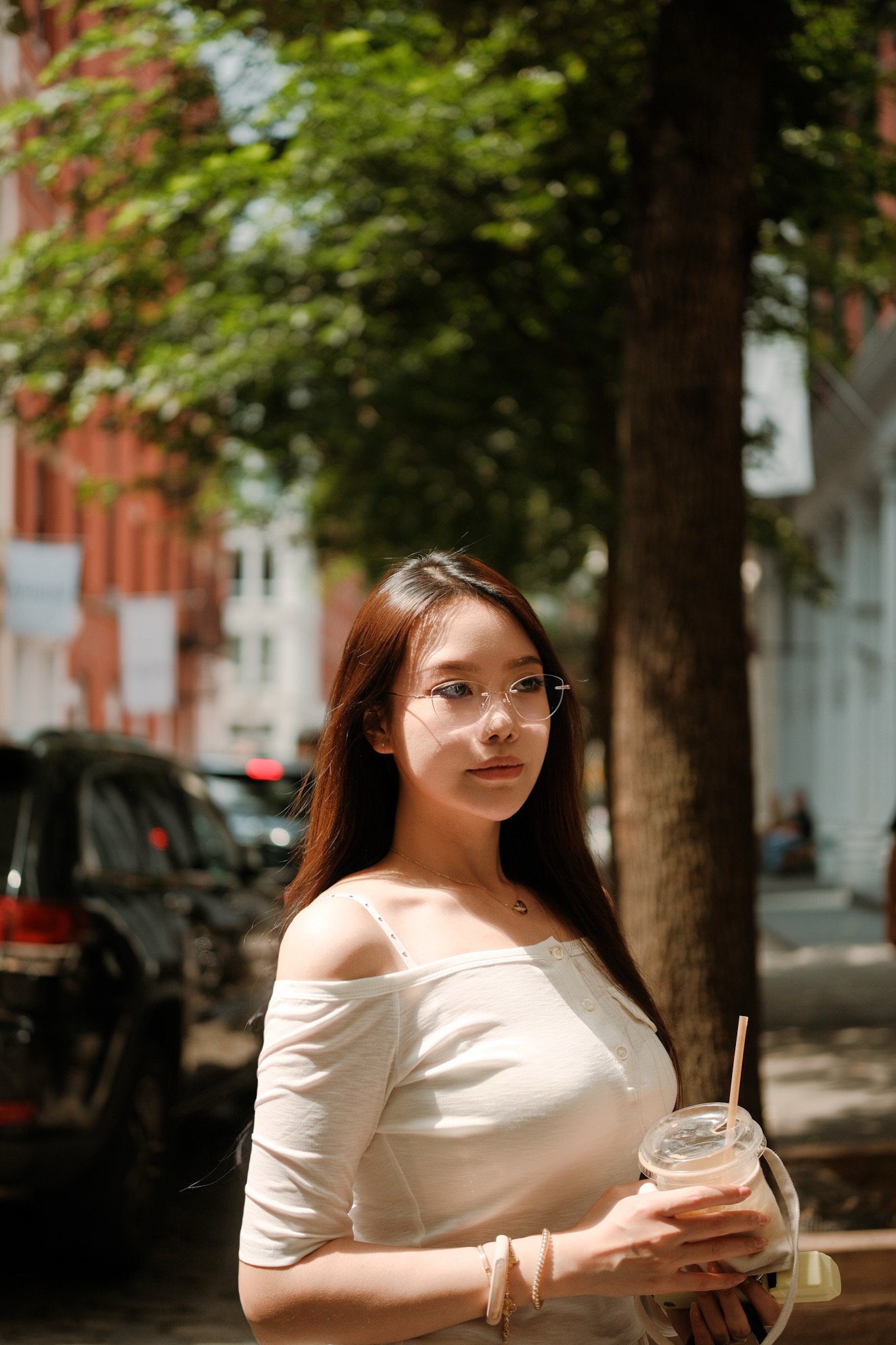 A young woman with long brown hair and glasses standing outdoors on a sunny day, holding a drink in a clear plastic cup with a straw, wearing a white off-shoulder top, trees and parked cars in the background.