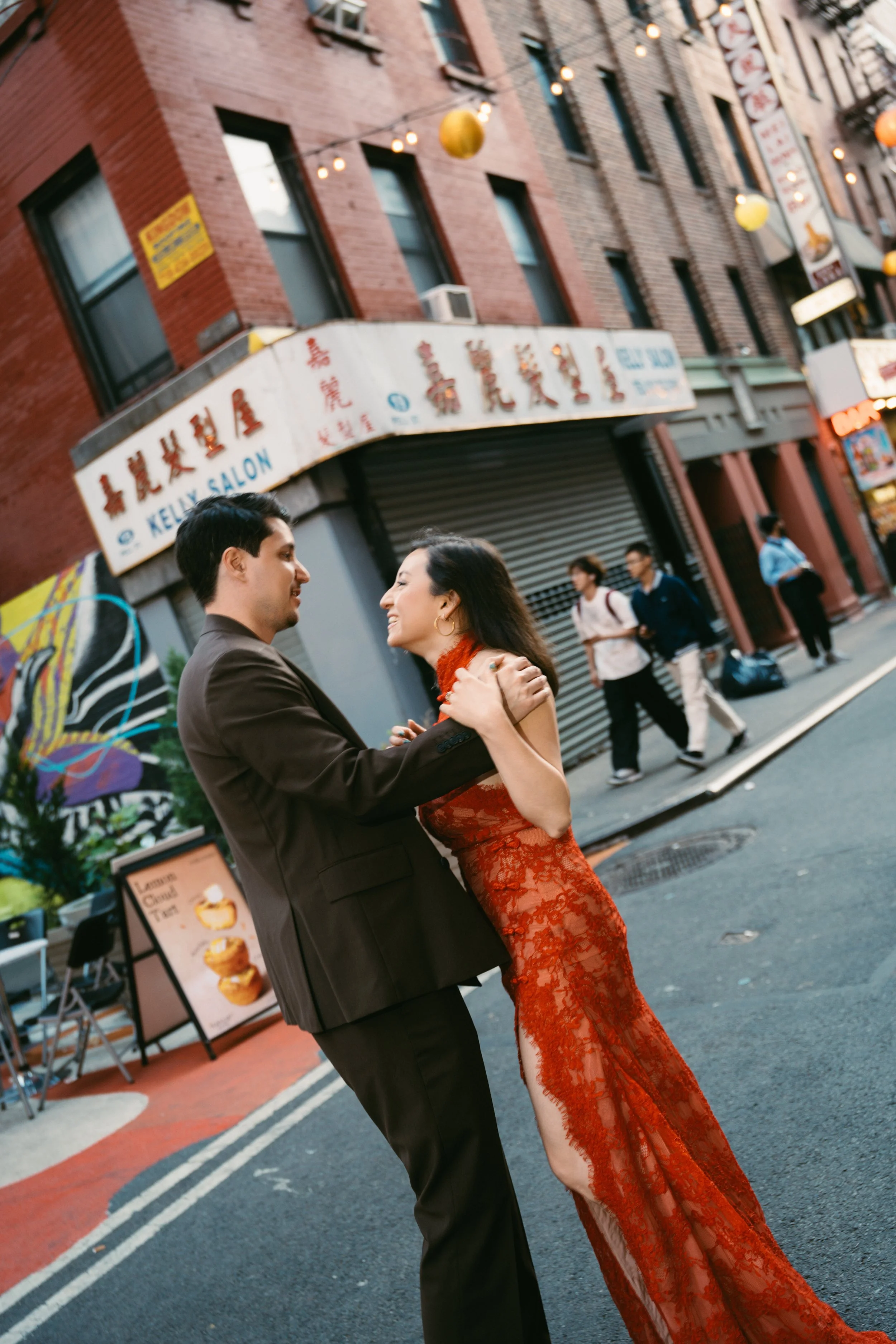 A man and woman dance closely on a city street at sunset, with people walking and storefronts behind them, including a salon and various signs and lanterns.