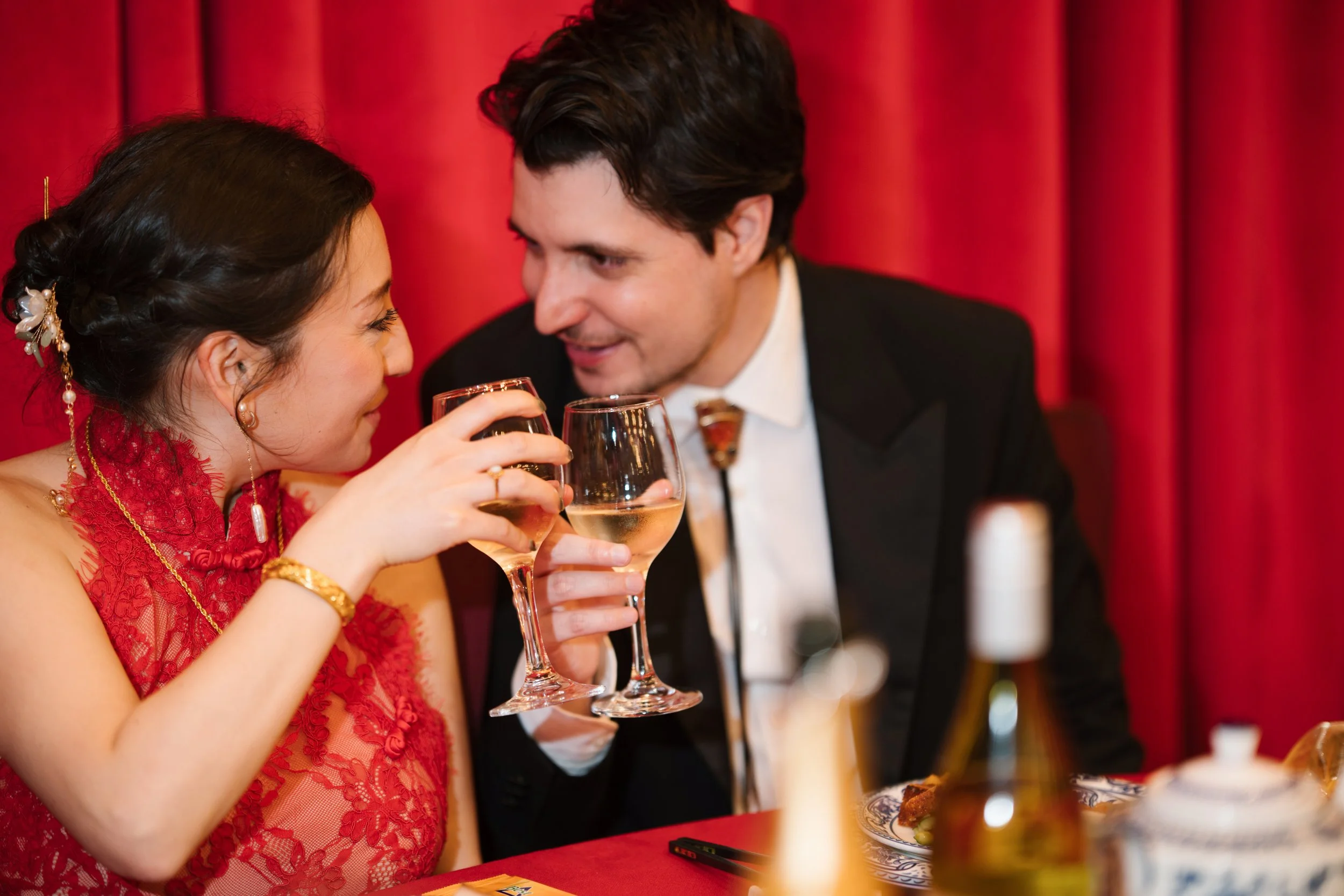 A man and woman with light skin dressed formally, toasting with glasses of white wine at an elegant event with a red curtain backdrop.