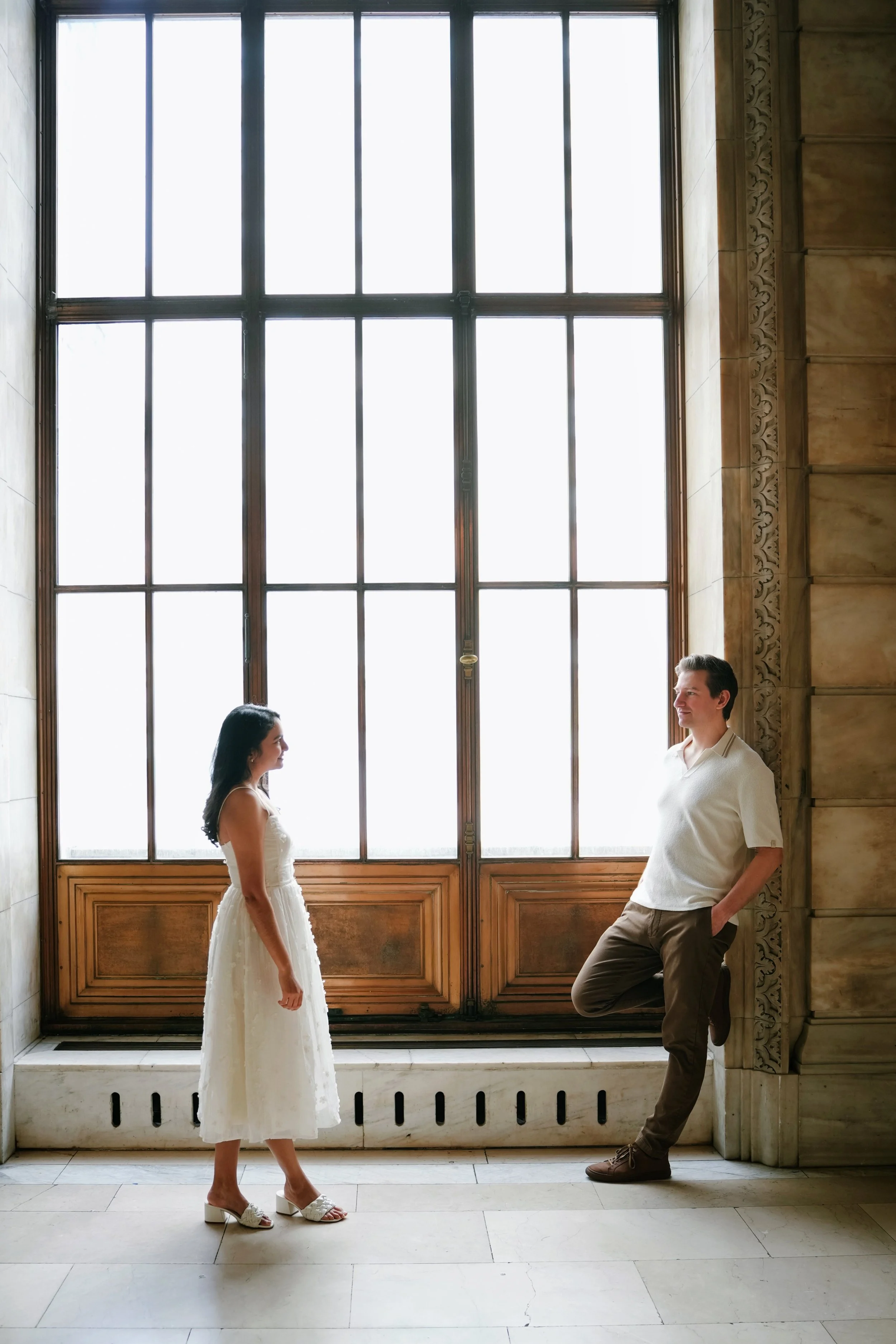 A woman in a white dress and high-heeled shoes standing and facing a man in casual clothes who is leaning against a wall near a large window with decorative wood details, in a historic or elegant building.
