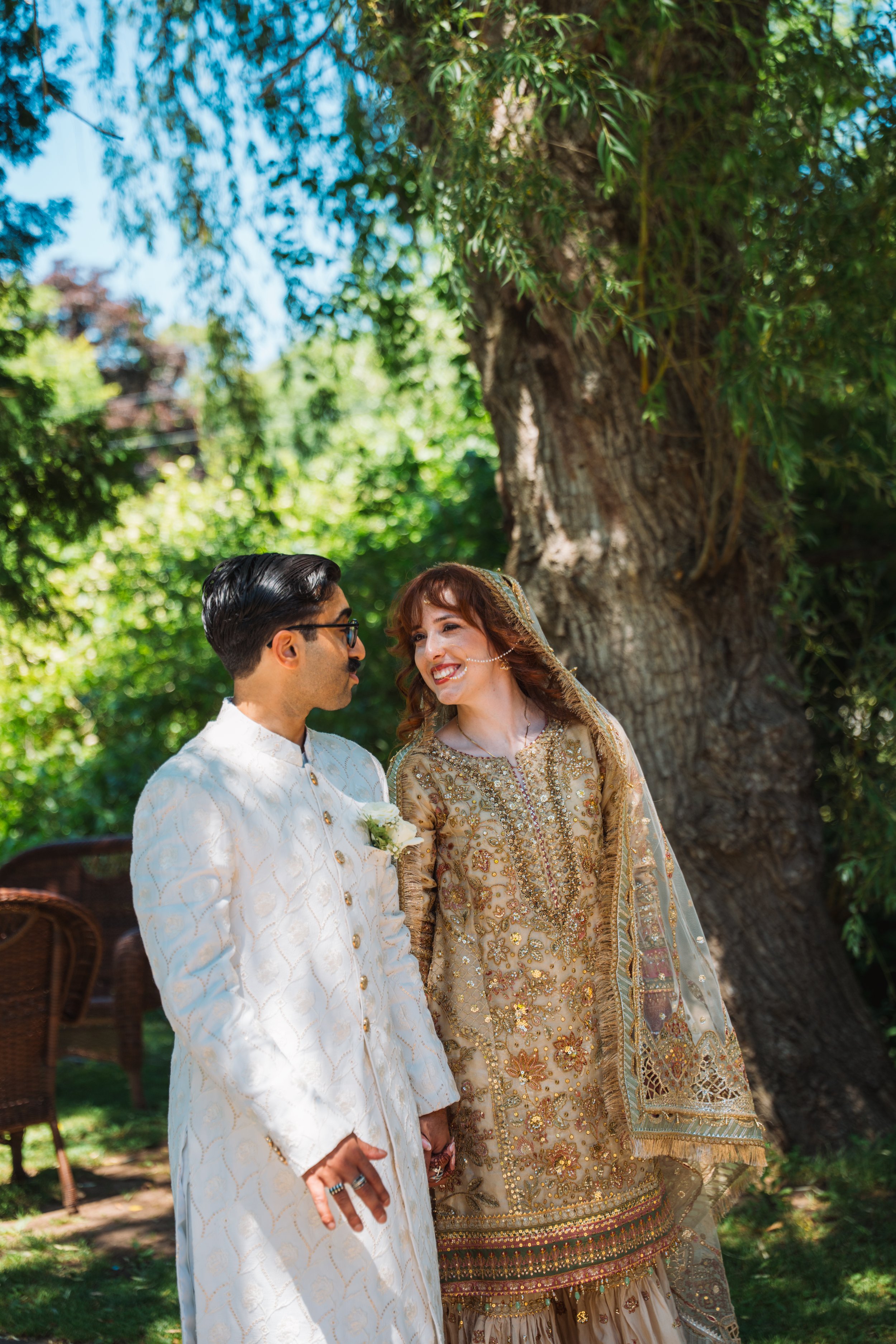 A man and woman dressed in wedding attire standing outdoors under a large tree, holding hands and smiling at each other.